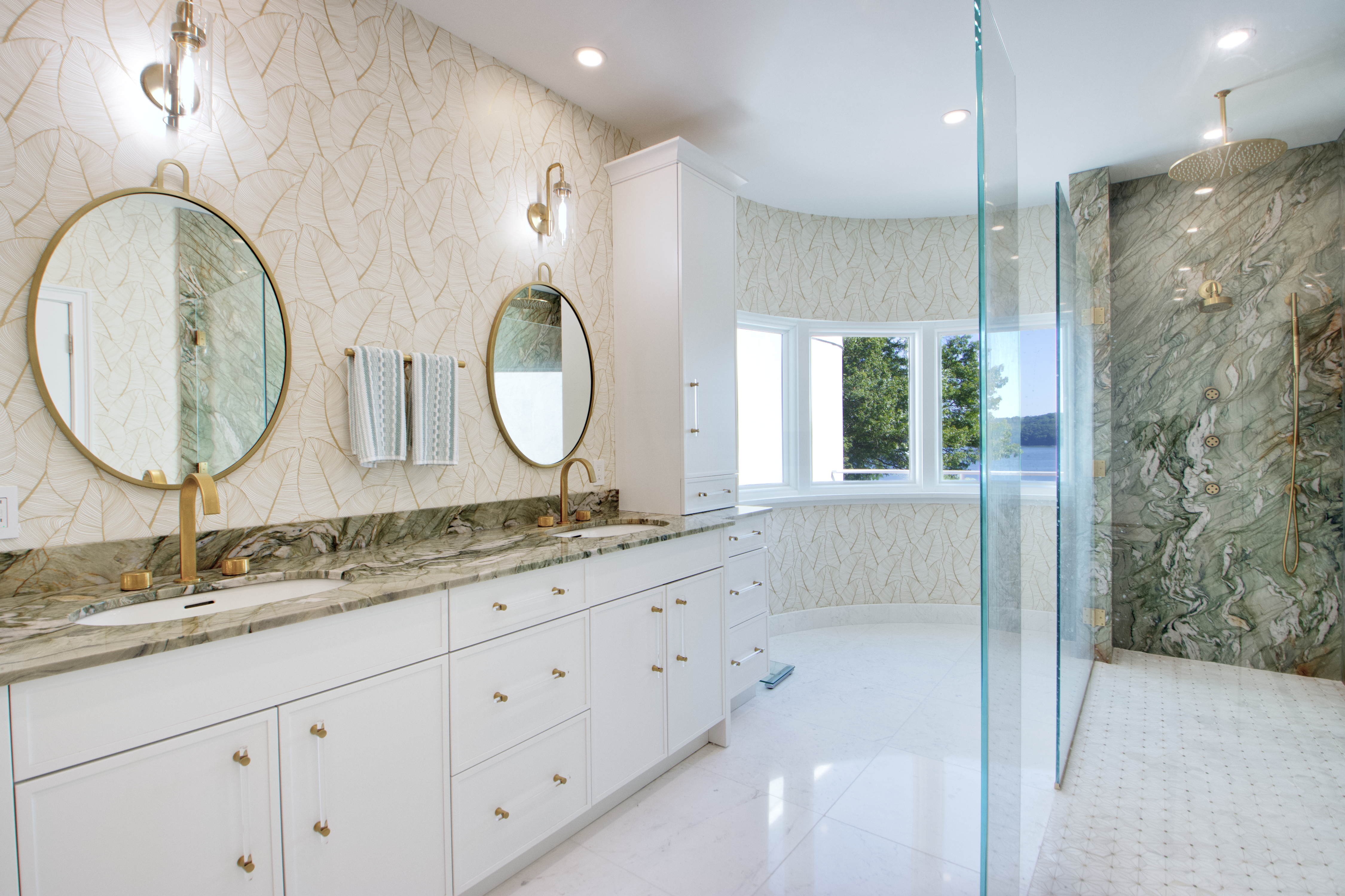 This luxurious primary bathroom features a double vanity with white cabinetry, gold hardware, and a striking green marble countertop. A large glass-enclosed walk-in shower with matching marble wall tiles and gold fixtures sits to the right, while a curved bay window provides natural light and a scenic view. The elegant wallpaper and polished flooring create a sophisticated, high-end atmosphere.