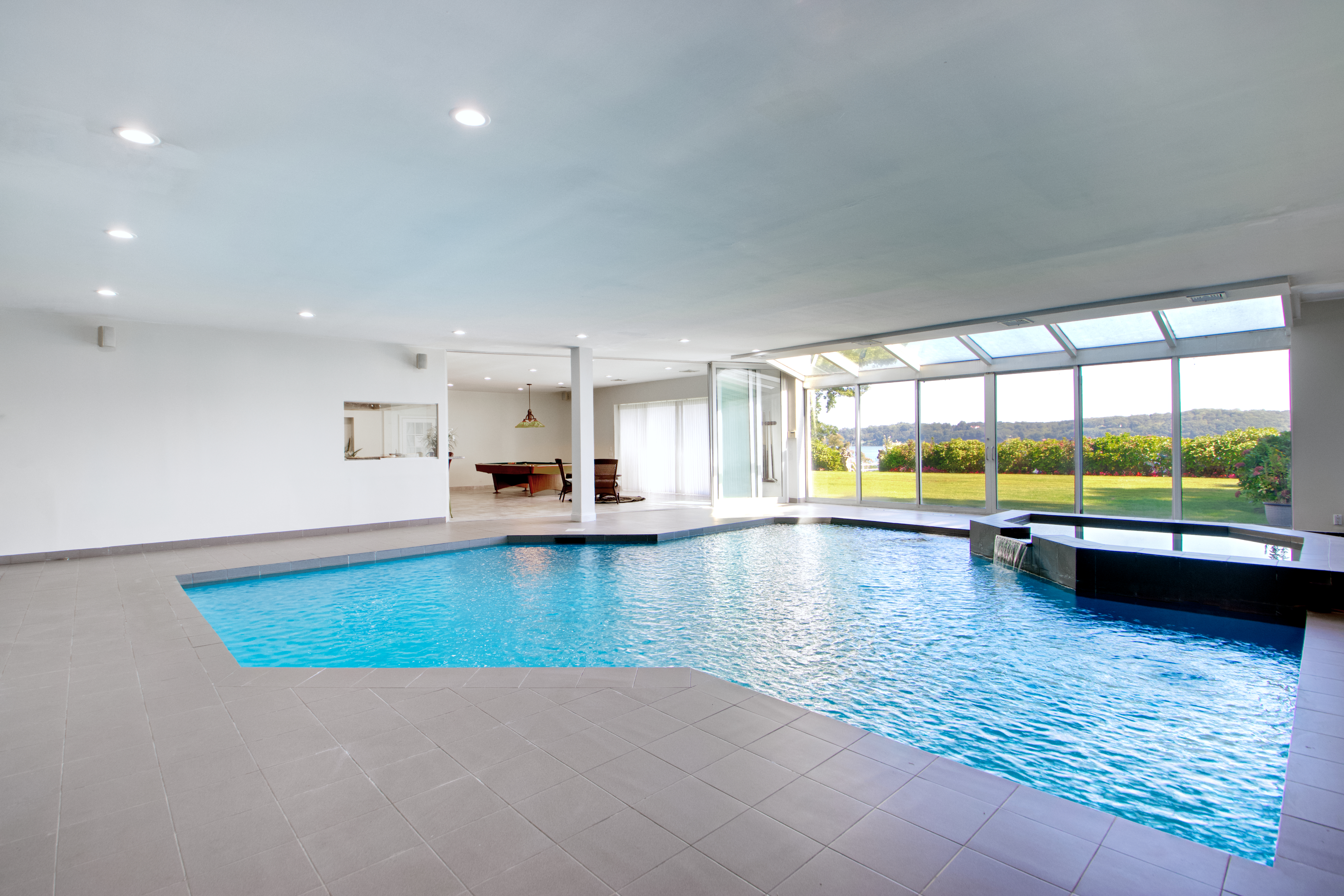 This expansive indoor pool room features a large, irregularly shaped swimming pool with an integrated spa, surrounded by neutral grey tiling. A wall of floor-to-ceiling glass windows and a glass-paneled ceiling provide abundant natural light and a seamless view of the lush green lawn and waterfront beyond. The space transitions into an adjacent room containing a pool table, creating a versatile and luxurious entertainment area.