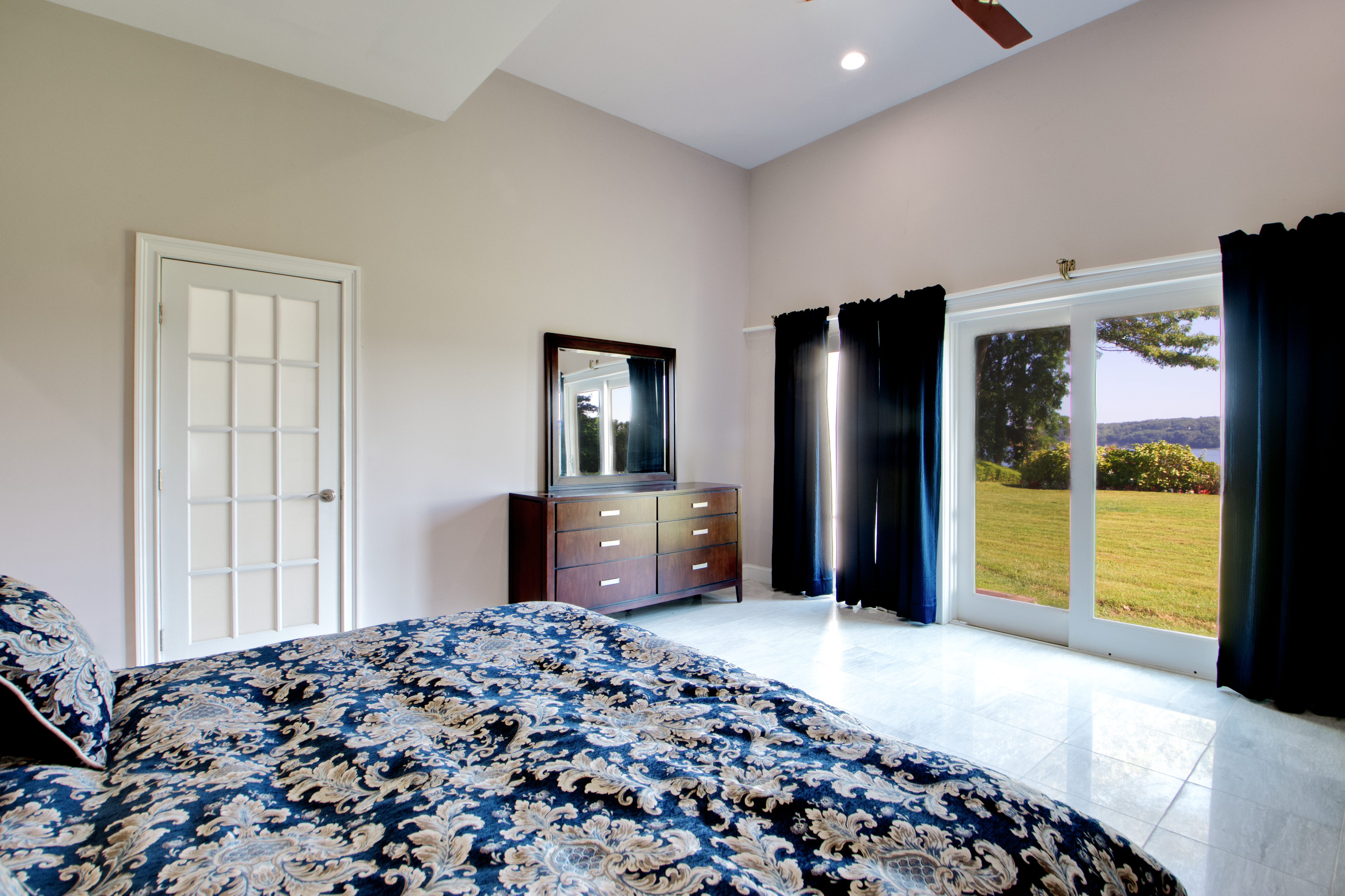 This spacious primary bedroom features high vaulted ceilings and polished tile flooring, creating an airy and elegant atmosphere. A large sliding glass door provides abundant natural light and a scenic view of the lush green backyard, while a dark wood dresser with a matching mirror adds a touch of traditional warmth. The room is anchored by a bed with a bold, ornate blue and gold patterned comforter, emphasizing a classic and comfortable aesthetic.