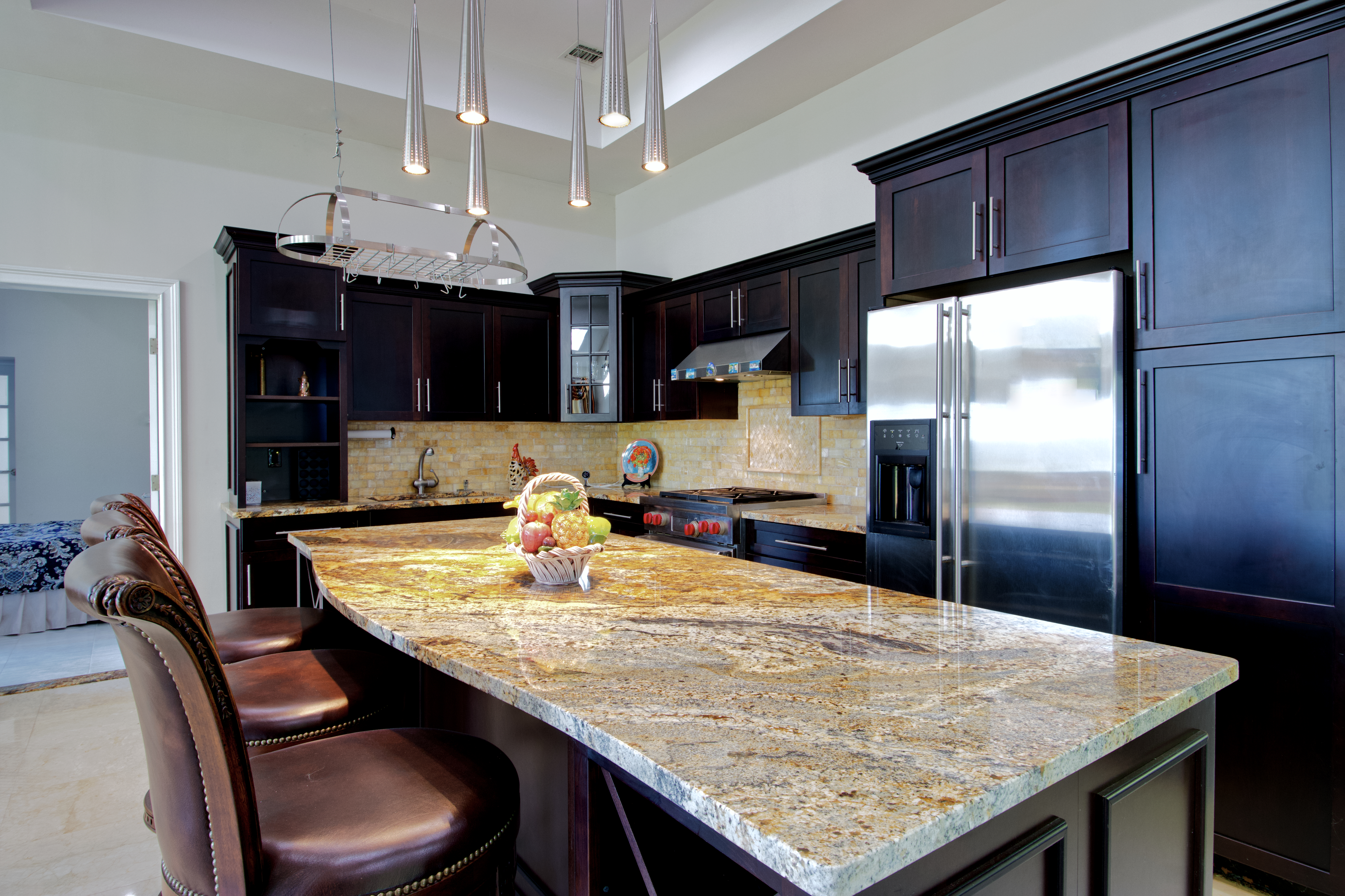 This elegant kitchen features dark wood cabinetry contrasted by a large, expansive granite island that serves as the centerpiece of the room. The space is illuminated by modern, slender pendant lights hanging above the island, while stainless steel appliances and a tiled backsplash add a touch of sophistication. The perspective is a wide-angle shot from the island, showcasing the functional layout and the warm, inviting atmosphere of the home.