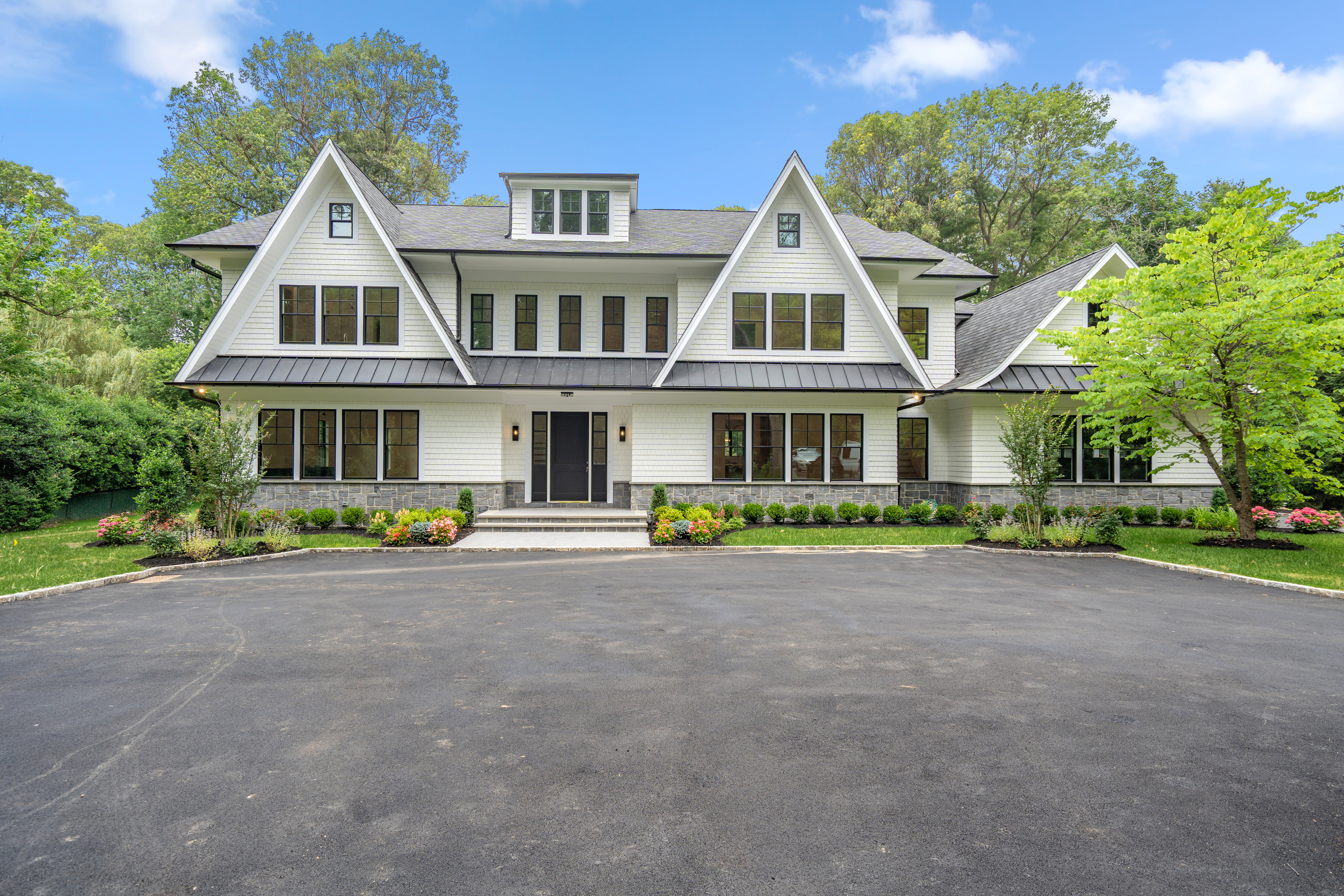 This stunning, newly constructed modern farmhouse features a crisp white exterior with dark-framed windows and a contrasting dark metal roof. The symmetrical facade is complemented by a stone base, a manicured garden bed, and a wide, paved driveway leading to a grand central entryway. The cinematic perspective captures the home's impressive scale and elegant curb appeal against a bright blue sky.