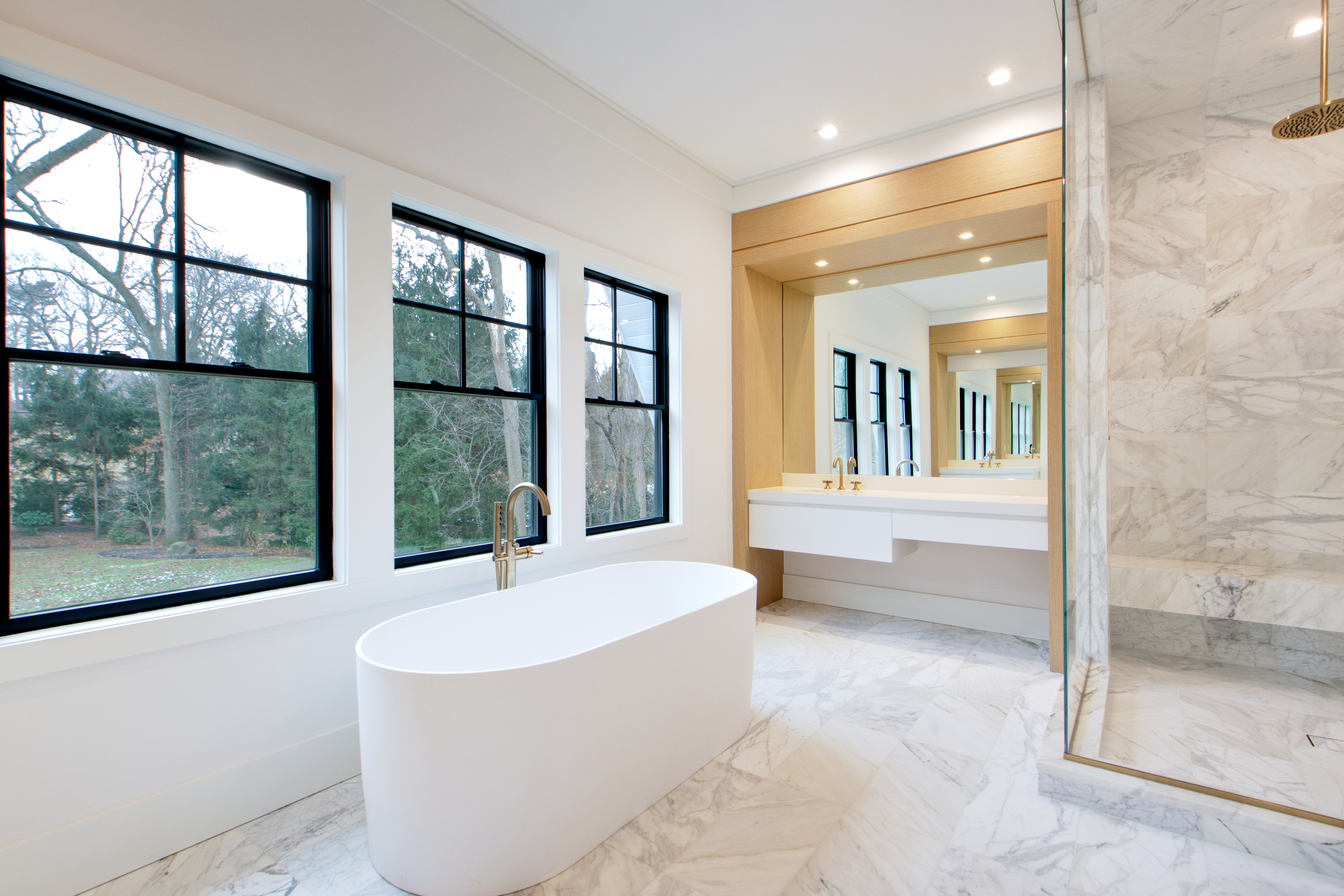 This luxurious primary bathroom features a sleek, freestanding white soaking tub positioned beneath large windows that offer a serene view of the outdoors. The space is anchored by a floating vanity with warm wood accents and a large mirror, complemented by a spacious walk-in marble shower. The overall aesthetic is modern and sophisticated, utilizing high-end materials like marble flooring and gold-toned fixtures to create a spa-like atmosphere.