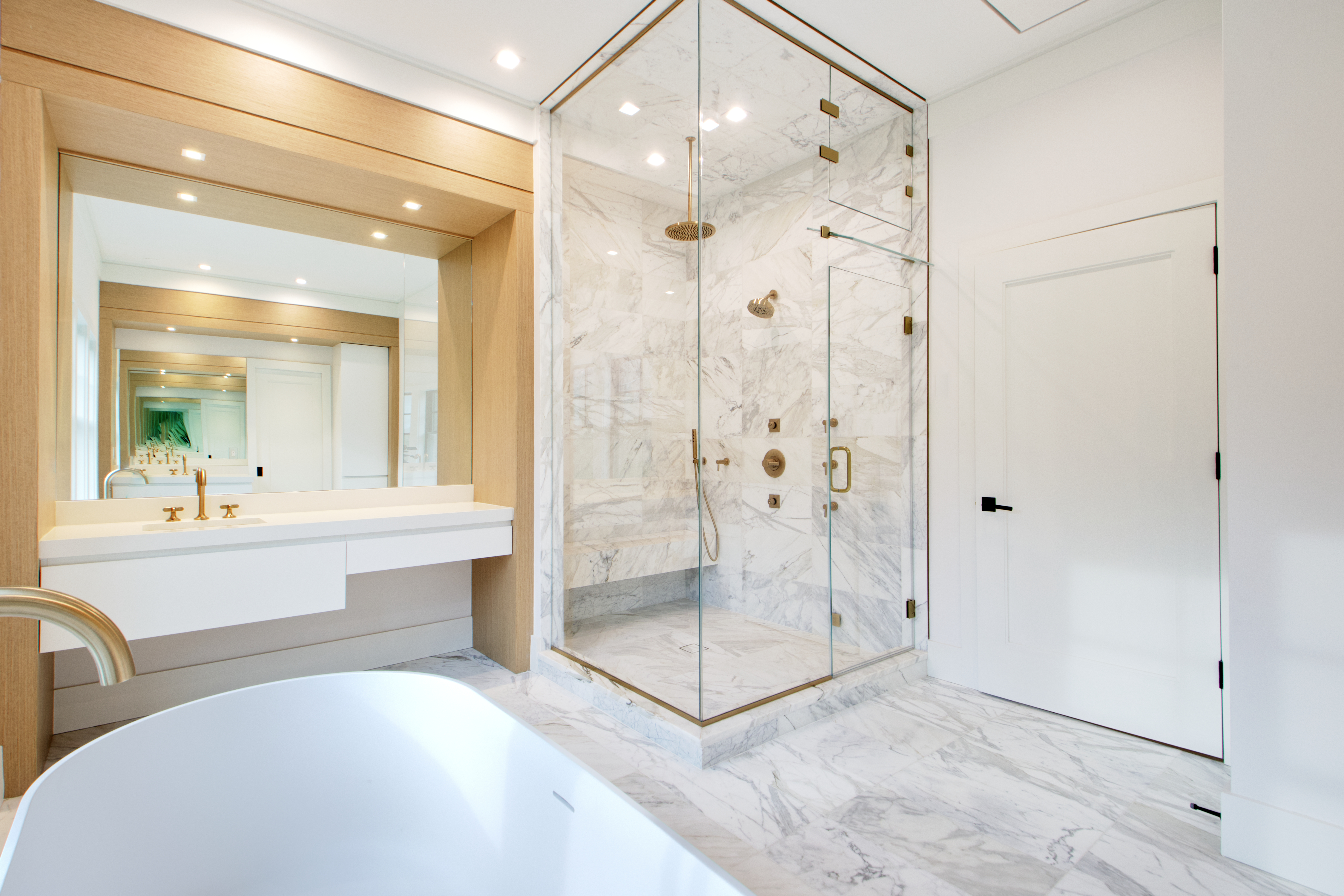This luxurious primary bathroom features a sleek, modern aesthetic with high-end marble tiling throughout the floors and shower enclosure. A prominent glass-enclosed walk-in shower with brass fixtures stands beside a minimalist white vanity and a deep soaking tub, creating a spa-like atmosphere. The warm wood accents around the mirror add a touch of organic elegance to the bright, clean space.