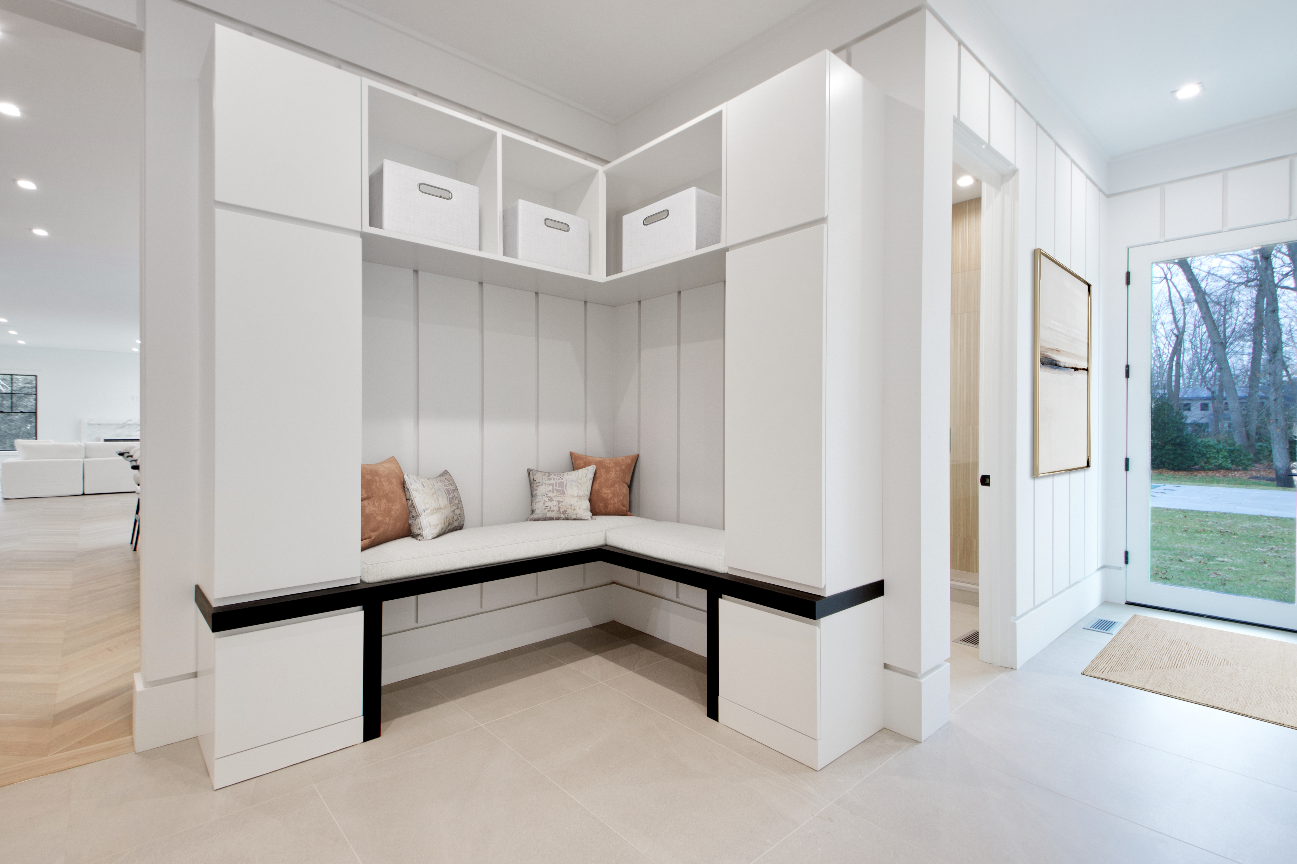 This bright and modern entryway features a custom built-in mudroom bench with white cabinetry, open storage cubbies, and a comfortable cushioned seat. The space is accented by a dark wood trim, decorative throw pillows, and vertical shiplap wall paneling that adds texture and depth. A glass-paned door allows natural light to flood the area, creating an inviting and organized transition space for the home.