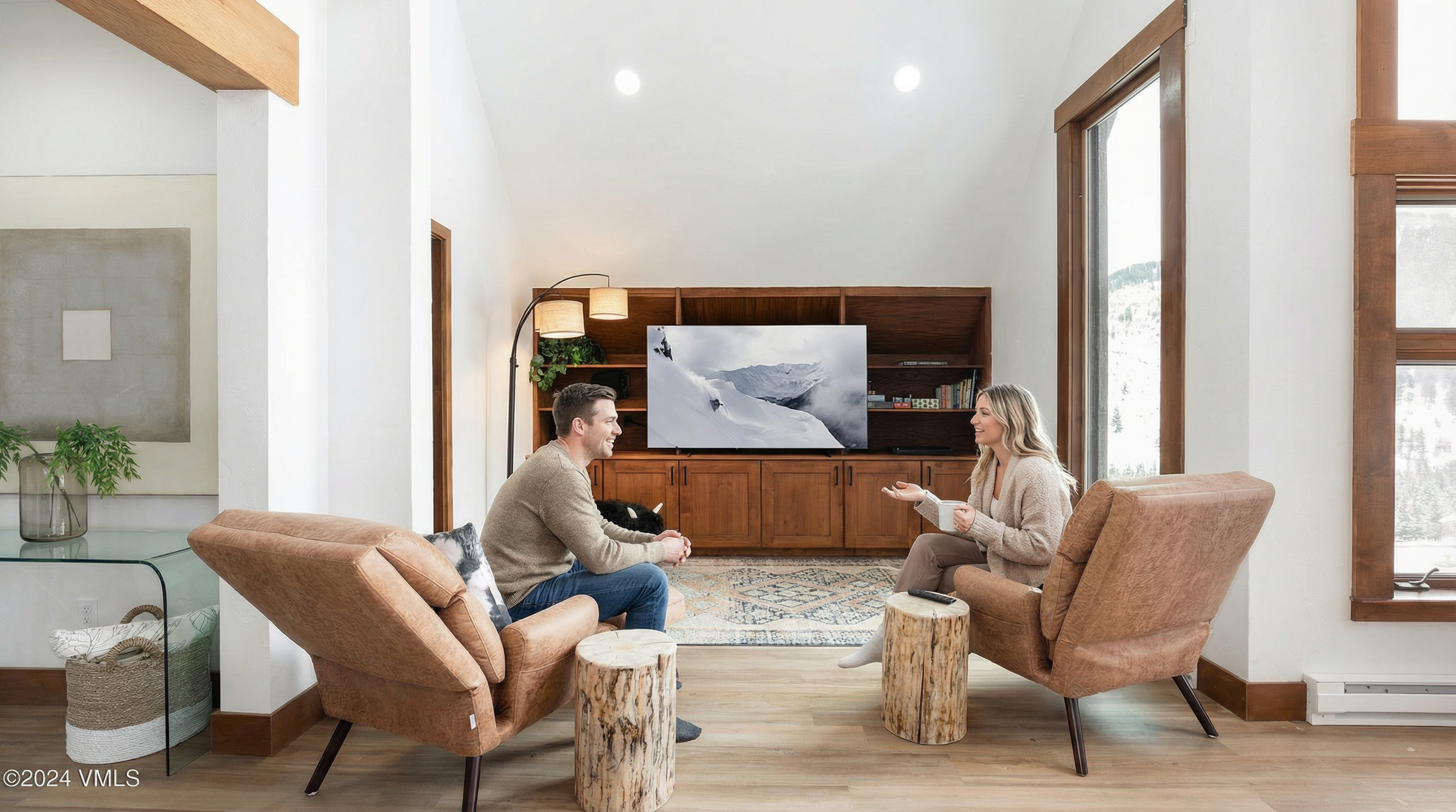 This is an interior shot of a living room featuring a man and woman sitting in stylish, caramel-colored chairs with a wooden stump serving as a side table beside each chair. The living room boasts a modern, open concept with a large built-in wooden entertainment center. Natural light floods in through a large window, complementing the light hardwood floors and neutral color palette, creating a warm and inviting atmosphere.