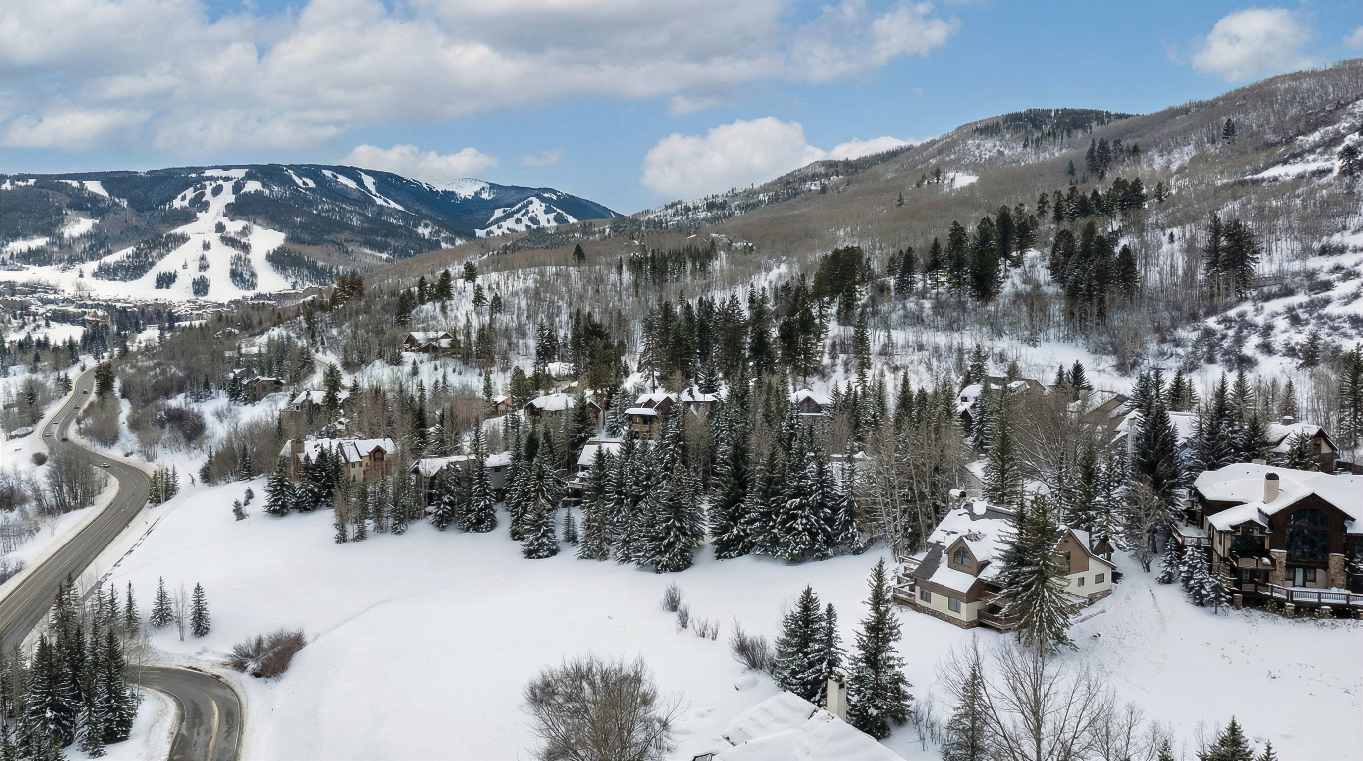 This aerial view showcases a luxury mountain property in a snowy landscape. Several residences are visible, nestled among evergreen trees, with snow-covered roofs and exteriors. A winding road leads into the neighborhood, offering access to the properties and demonstrating the serene, natural setting.