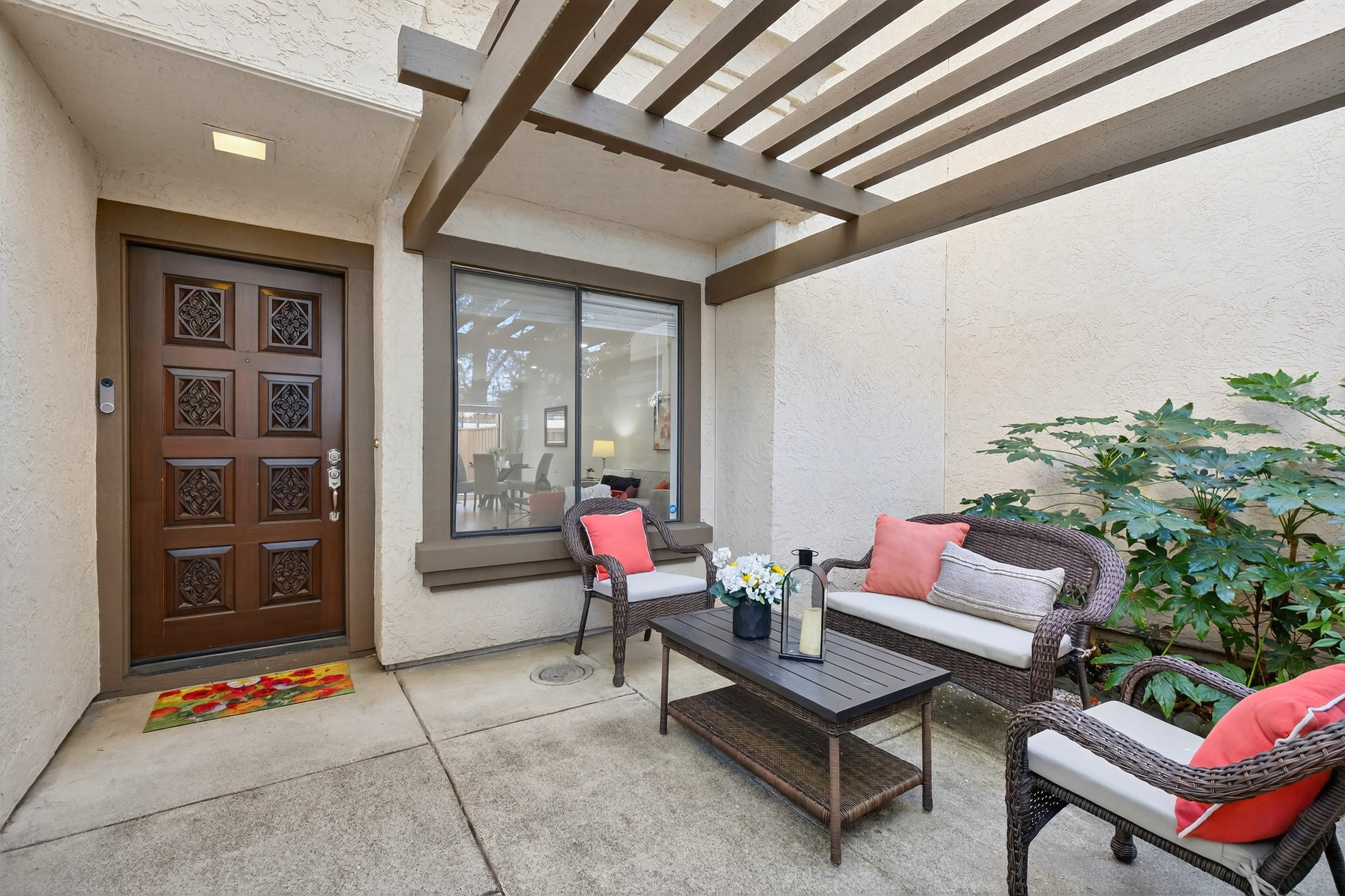 This inviting covered patio features a dark wood pergola overhead and a set of wicker-style outdoor furniture with plush cushions and vibrant coral-colored pillows. A dark coffee table sits in the center, complemented by a decorative lantern and a small floral arrangement, creating a cozy seating area adjacent to the home's front entry. The space is framed by a textured stucco wall and a large window that offers a glimpse into the interior living space, providing a warm and welcoming first impression.
