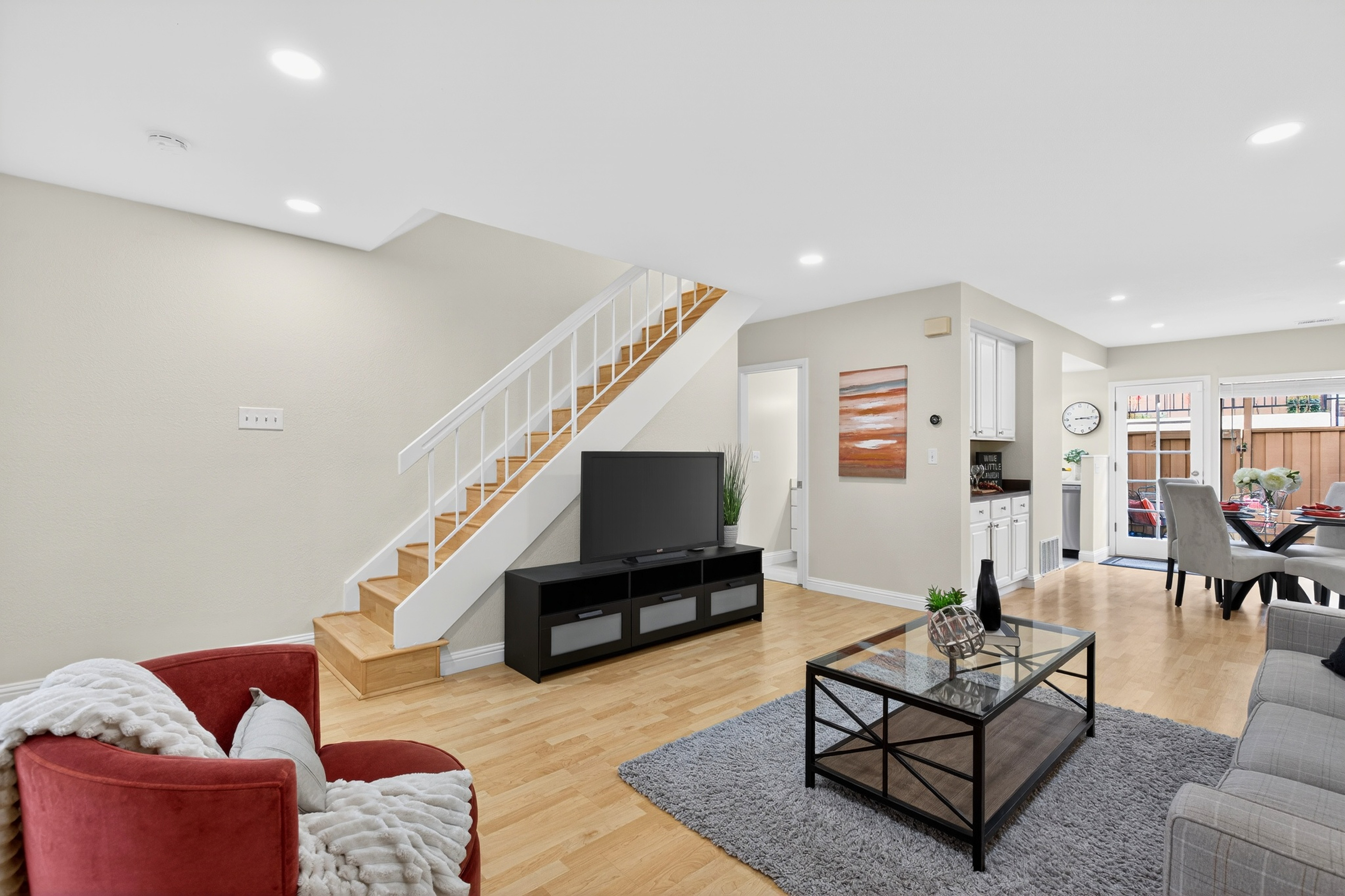 This open-concept living room features light wood flooring and a neutral color palette, creating a bright and inviting atmosphere. The space includes a staircase with white railings, a black media console supporting a television, and a glass-topped coffee table centered on a textured gray area rug. A red armchair adds a pop of color, while the background reveals a dining area and a glass door leading to an outdoor space.