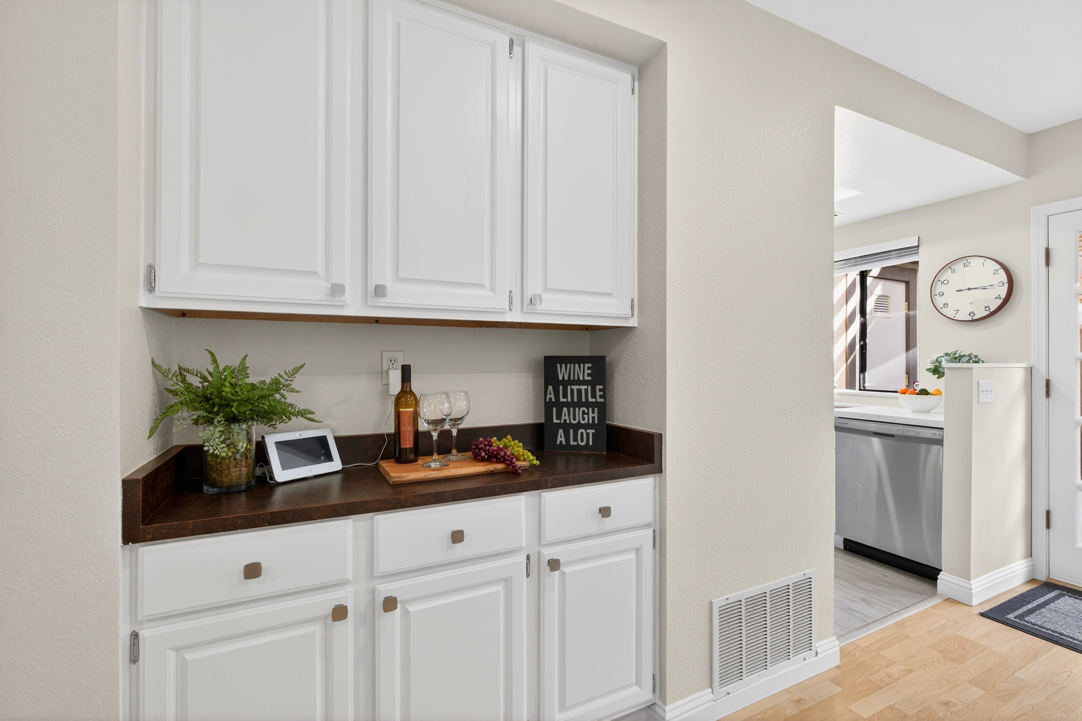 This image showcases a functional kitchen wet bar or butler's pantry area featuring crisp white cabinetry paired with a dark, textured countertop. The space is styled with a decorative sign, a small potted plant, and a wooden serving board with wine and grapes, creating an inviting atmosphere. A doorway to the right provides a glimpse into the main kitchen area, which includes a stainless steel dishwasher and a wall-mounted clock.