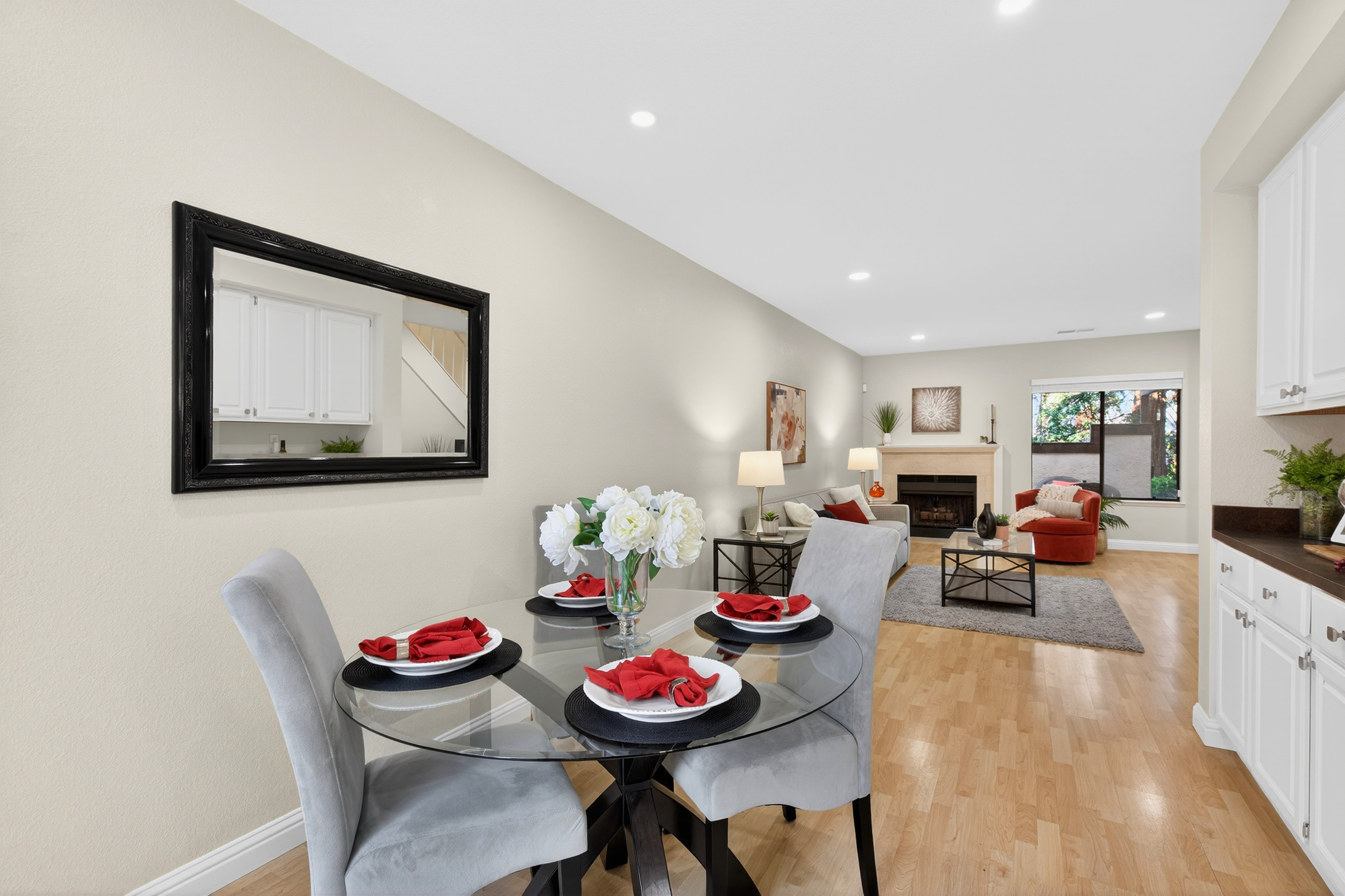 This open-concept living space features a bright dining area in the foreground with a glass-topped table and light gray upholstered chairs, accented by red napkins. The room flows seamlessly into a cozy living area with a fireplace, neutral-toned furniture, and light wood flooring throughout. A large mirror on the wall reflects the kitchen cabinetry, enhancing the sense of space and light in this inviting home.