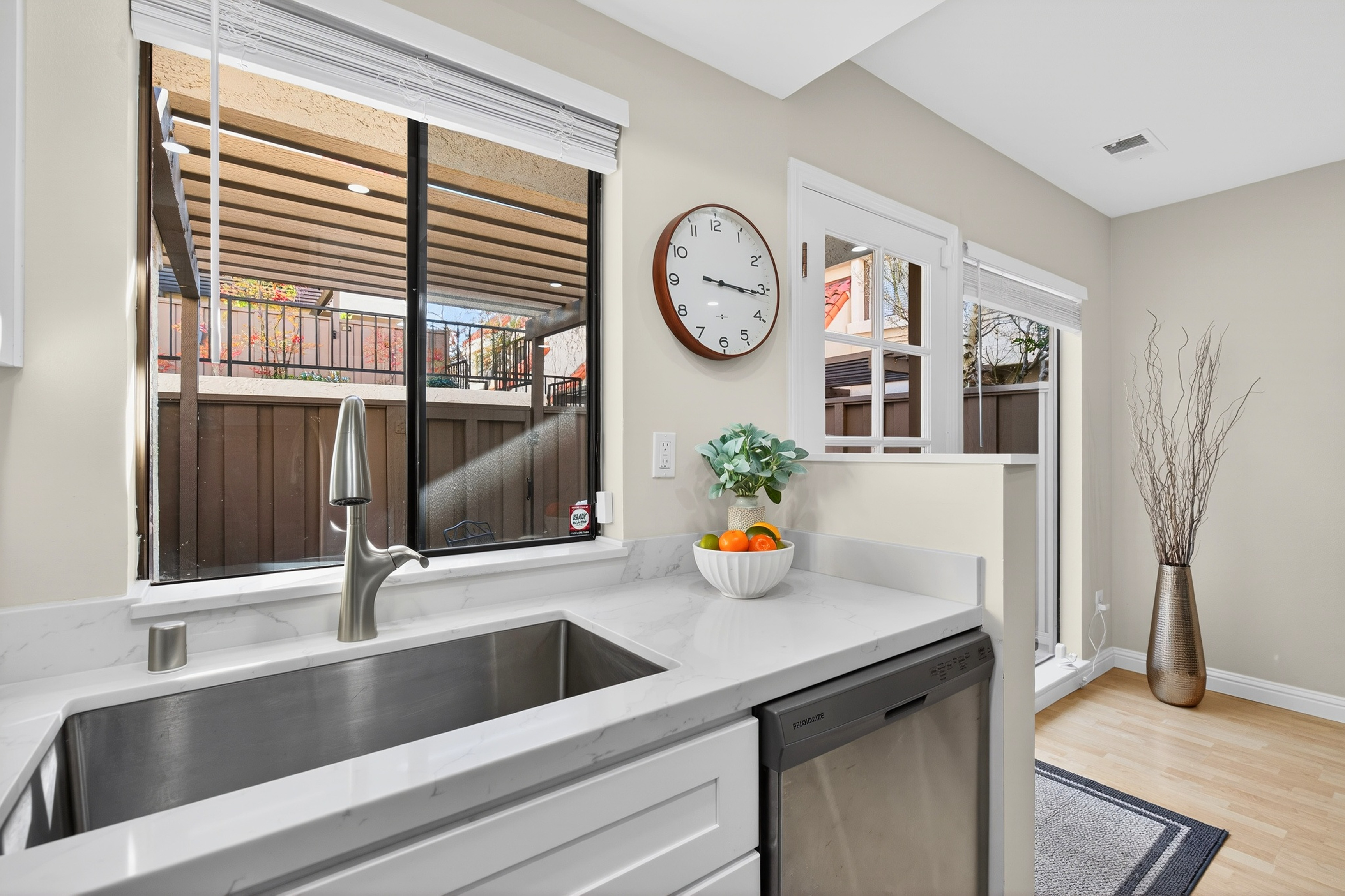 This bright kitchen area features a stainless steel sink set into a light-toned stone countertop with white cabinetry below. A large window above the sink provides a view of the outdoor patio, while a wall-mounted clock and a decorative bowl of fruit add a personal touch. The space transitions into an adjacent area with light wood flooring and a tall decorative vase, creating an open and inviting atmosphere.