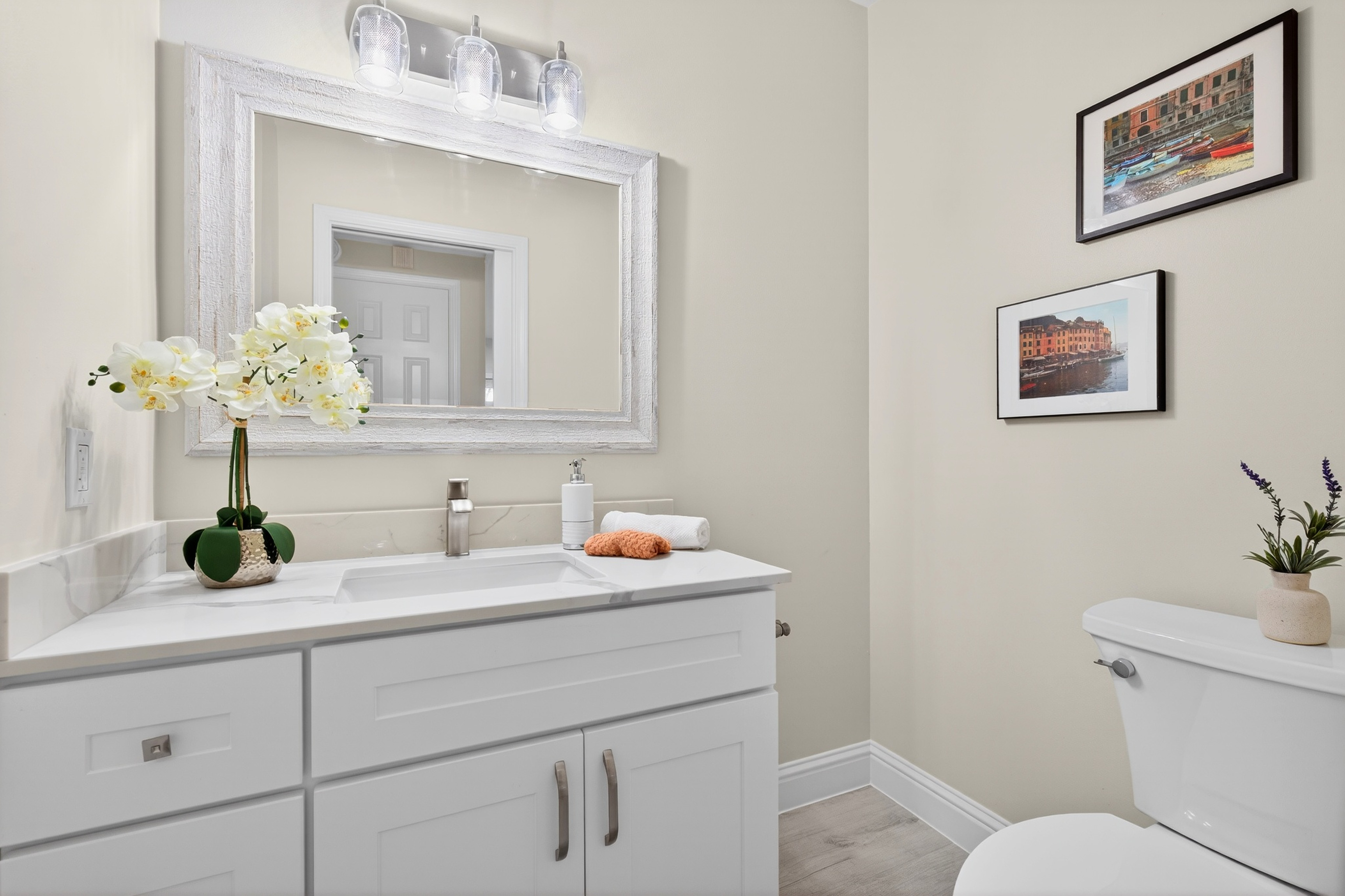 This bright and clean bathroom features a white vanity with a stone countertop, a rectangular undermount sink, and a decorative framed mirror. The space is accented with a floral arrangement, a soap dispenser, and two framed pictures on the wall, creating a welcoming and tidy atmosphere. The neutral color palette and simple fixtures provide a modern and functional aesthetic suitable for a guest bathroom.