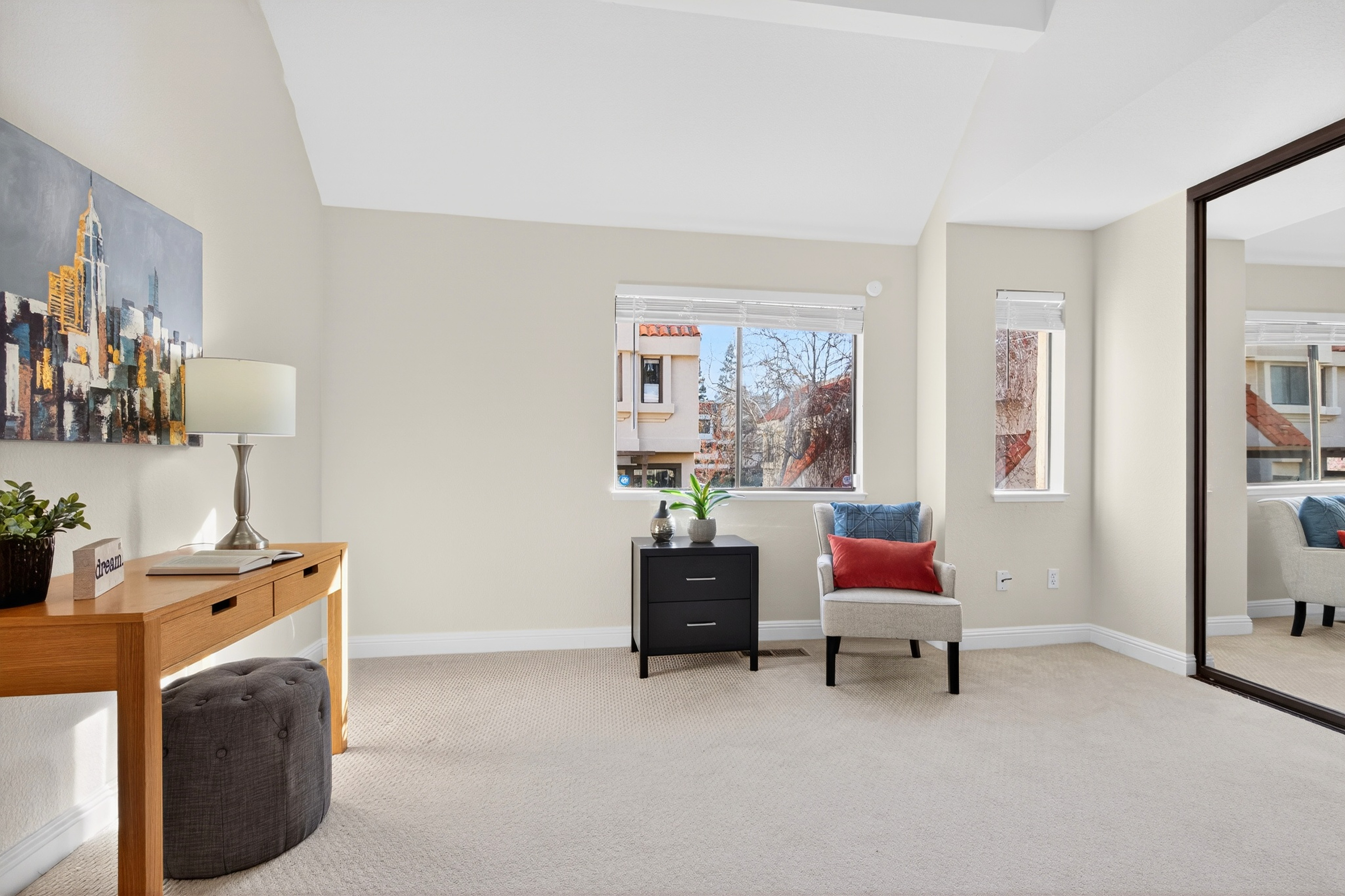 This bright and airy home office or study features neutral walls, plush carpeting, and a vaulted ceiling that enhances the sense of space. The room is furnished with a light wood desk, a dark grey ottoman, a black two-drawer nightstand, and a comfortable armchair accented with blue and red pillows. A large window and a smaller adjacent window provide natural light, while a large mirror on the right wall reflects the room's clean, minimalist aesthetic.