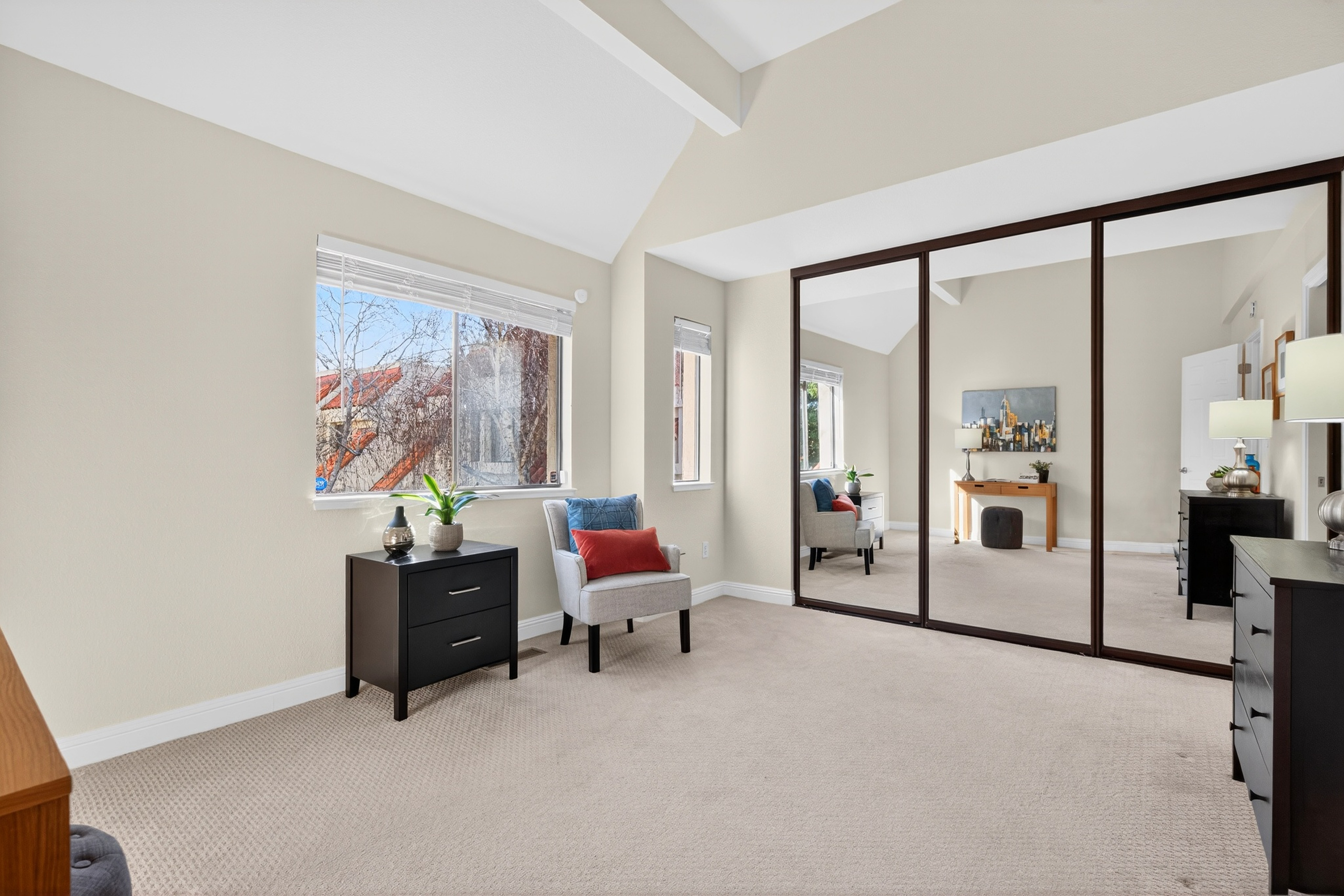 This bright bedroom features a vaulted ceiling and neutral wall tones, creating an airy and spacious atmosphere. The room is furnished with a small accent chair, a dark wood nightstand, and a large mirrored closet that reflects the opposite side of the room. A window provides natural light, while the carpeted flooring adds a comfortable, cozy feel to the space.