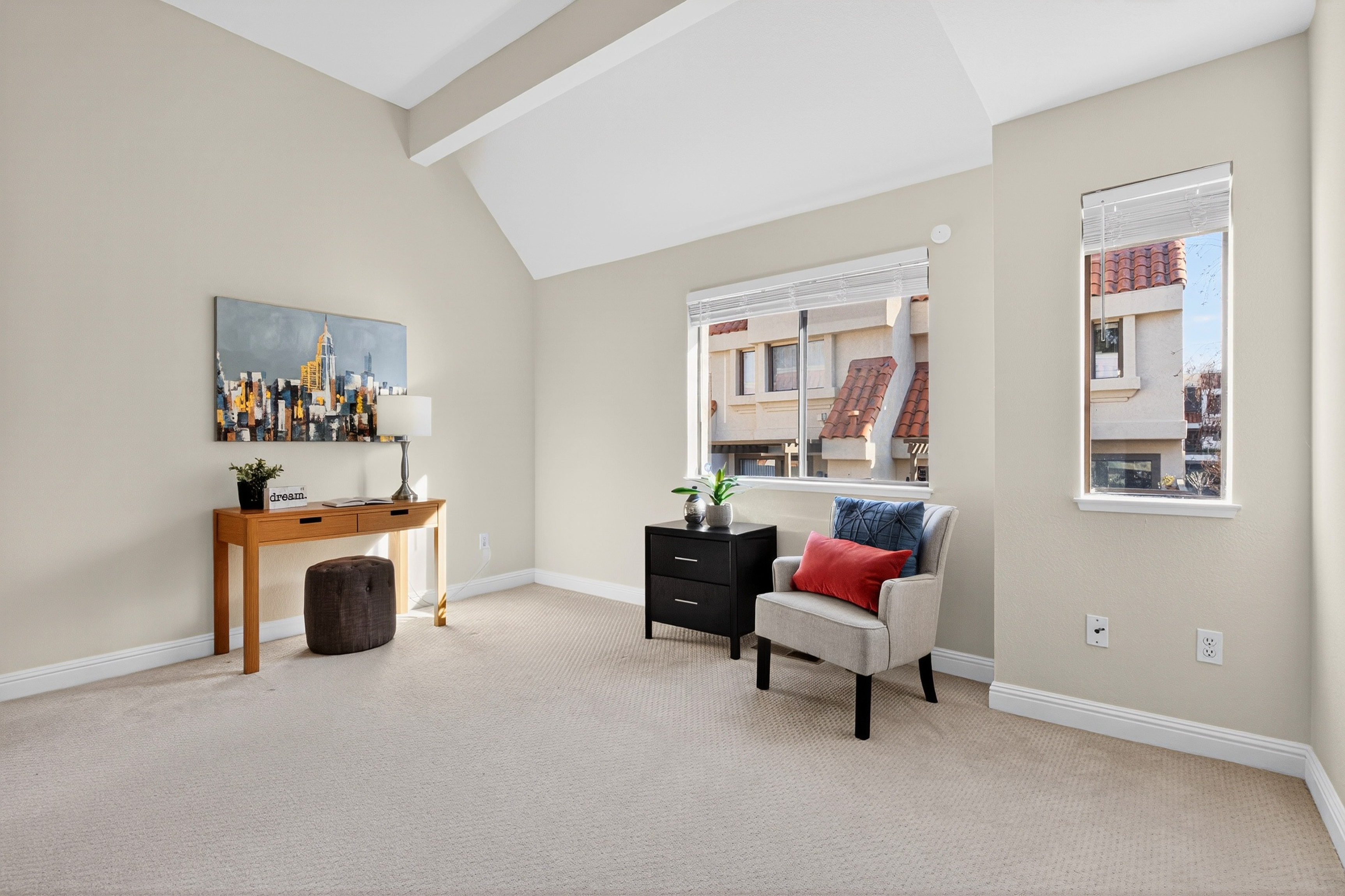 This bright and airy room features a vaulted ceiling and neutral wall tones, creating a versatile space suitable for a home office or reading nook. The room is furnished with a light wood desk, a dark storage cabinet, and a comfortable armchair accented with colorful pillows. Two windows provide natural light and a view of the neighboring residential architecture, while the carpeted flooring adds a soft, comfortable feel to the space.