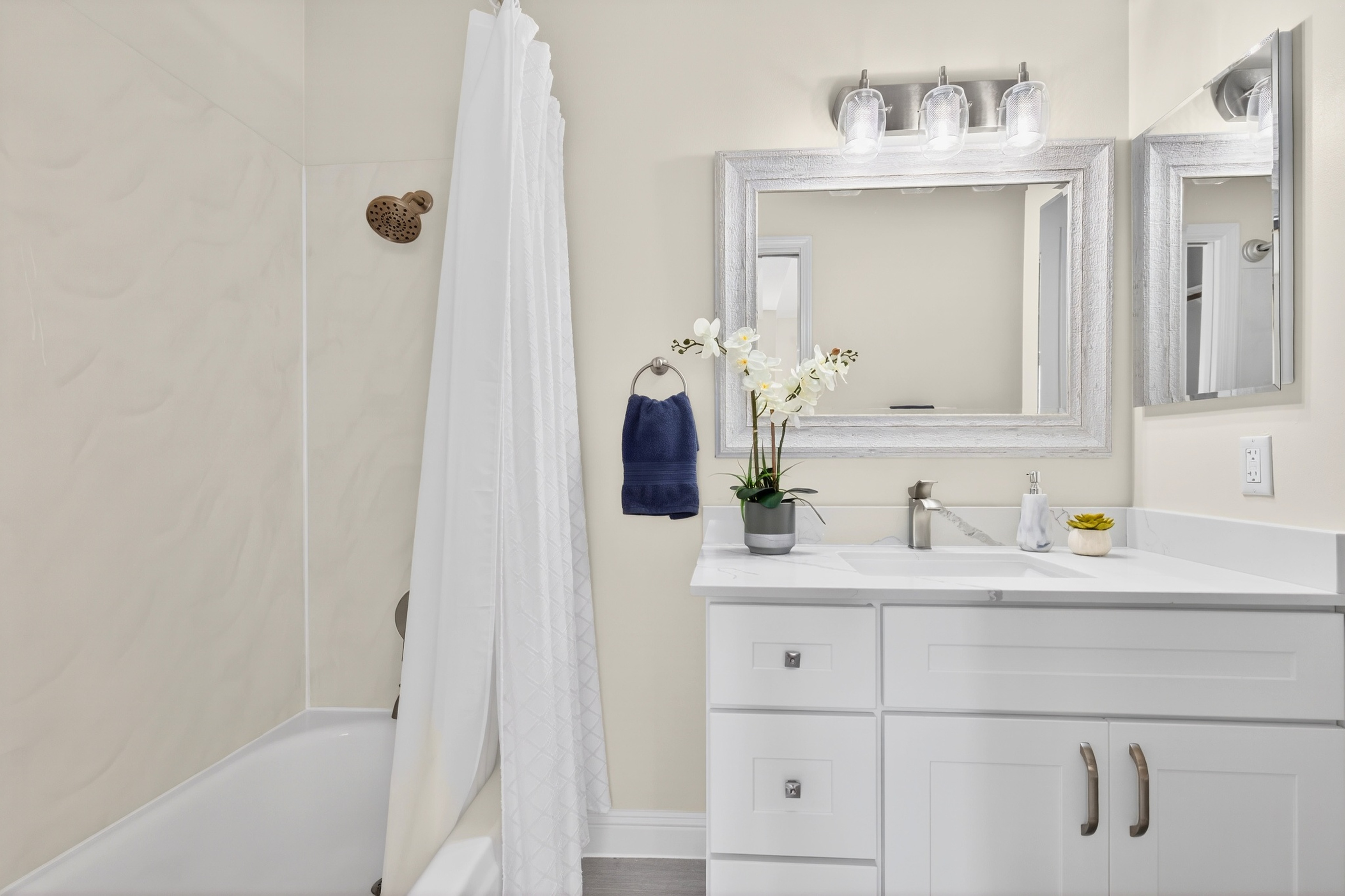 This bright and clean bathroom features a white vanity with a stone countertop and a rectangular undermount sink. A silver-framed mirror hangs above the vanity, complemented by a three-light fixture with glass shades. To the left, a shower-tub combination is enclosed by a white curtain, and a small potted orchid adds a touch of greenery to the counter.