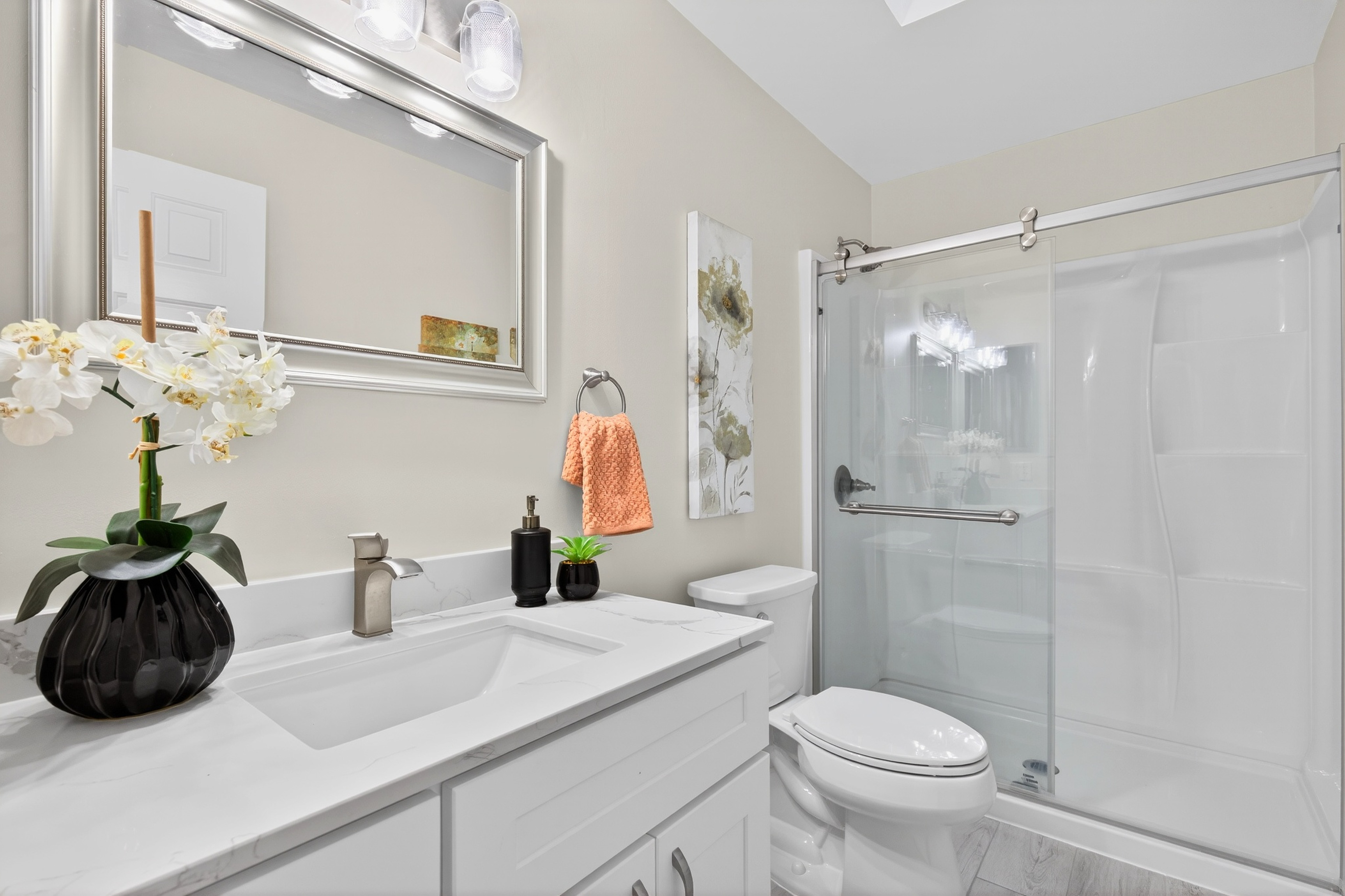 This bright, modern bathroom features a white vanity with a stone countertop, a silver-framed mirror, and a glass-enclosed shower stall. The space is accented with a decorative floral wall art piece, a peach-colored hand towel, and a small potted plant, creating a clean and inviting atmosphere. The perspective is a straight-on shot capturing the vanity, toilet, and shower in a cohesive, well-lit layout.