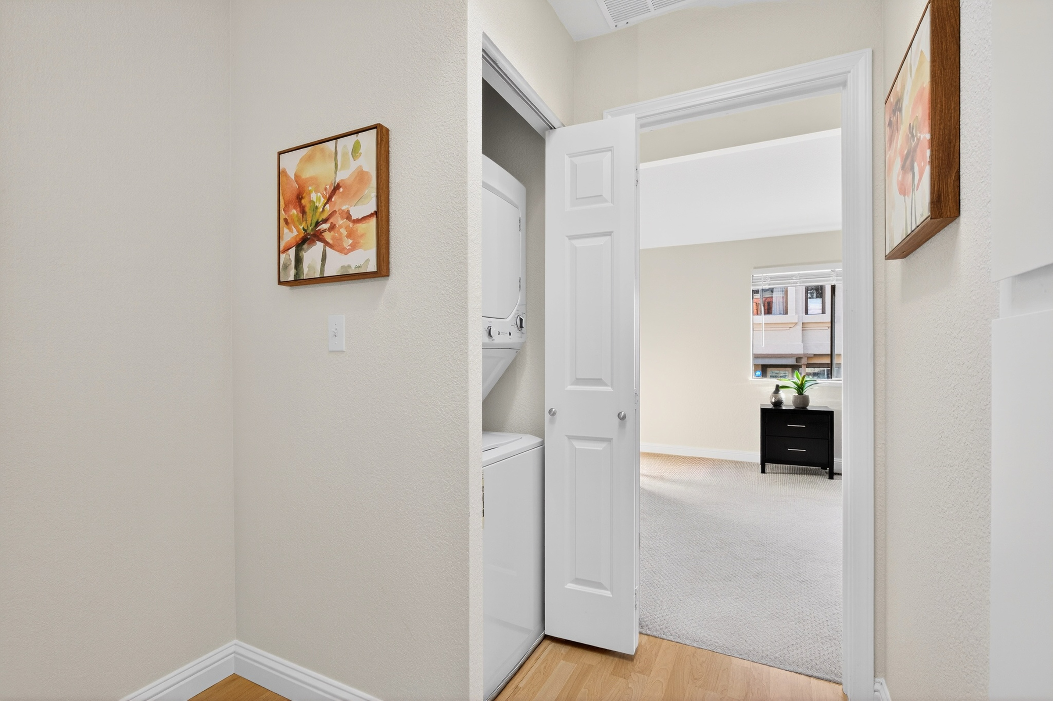 This image captures a laundry closet featuring a stacked washer and dryer unit, conveniently tucked away behind a white door. The foreground shows light-toned wood flooring, while the open door reveals a glimpse into a carpeted bedroom area with a black dresser and a window. The walls are painted a neutral tone, accented by framed floral artwork on either side of the doorway, creating a clean and functional transition space.