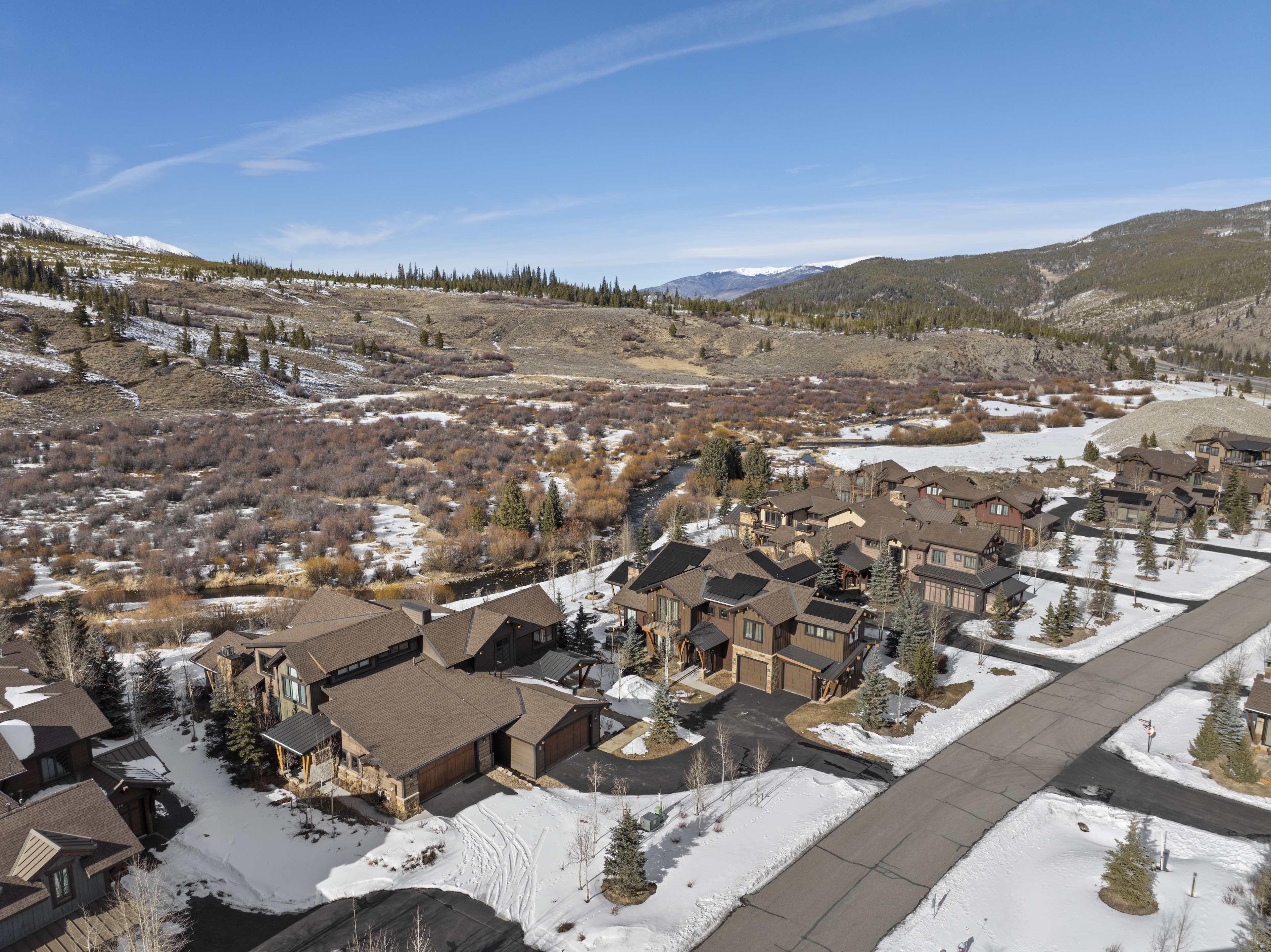 This is an aerial view of a residential neighborhood with upscale homes, showcasing the property's location and surrounding environment. The homes have a modern mountain aesthetic with brown tones and architectural details like stone accents and gabled roofs. Visible snowy patches suggest a desirable, possibly resort-like setting with scenic mountain views and a nearby river.