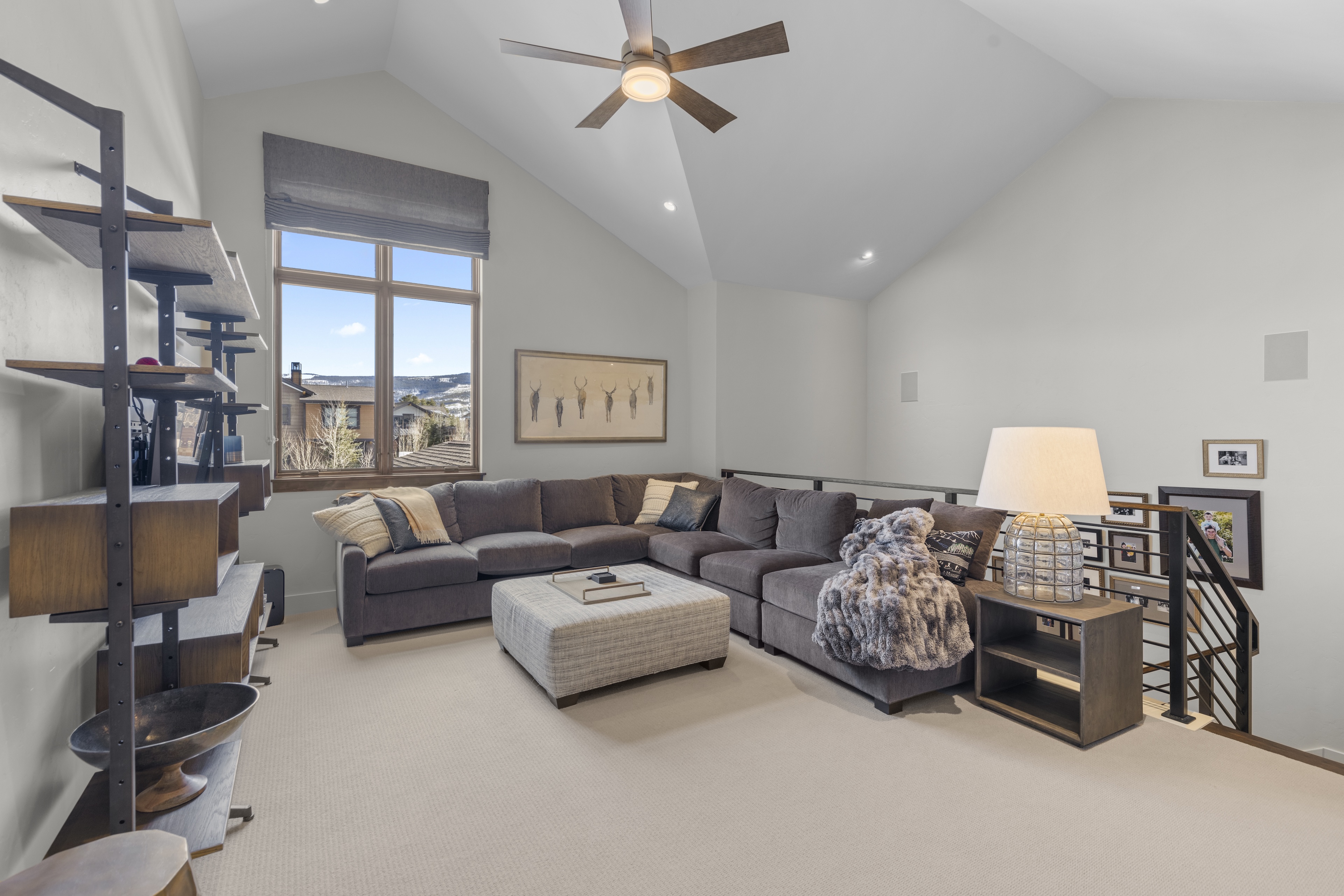 The image showcases a cozy living room with a large, sectional sofa and an ottoman that serves as a coffee table. A ceiling fan and recessed lighting illuminate the room, which features neutral tones and a large window offering a view of the outdoor landscape. Wooden shelves provide storage and adds a unique touch to the staging of the room.