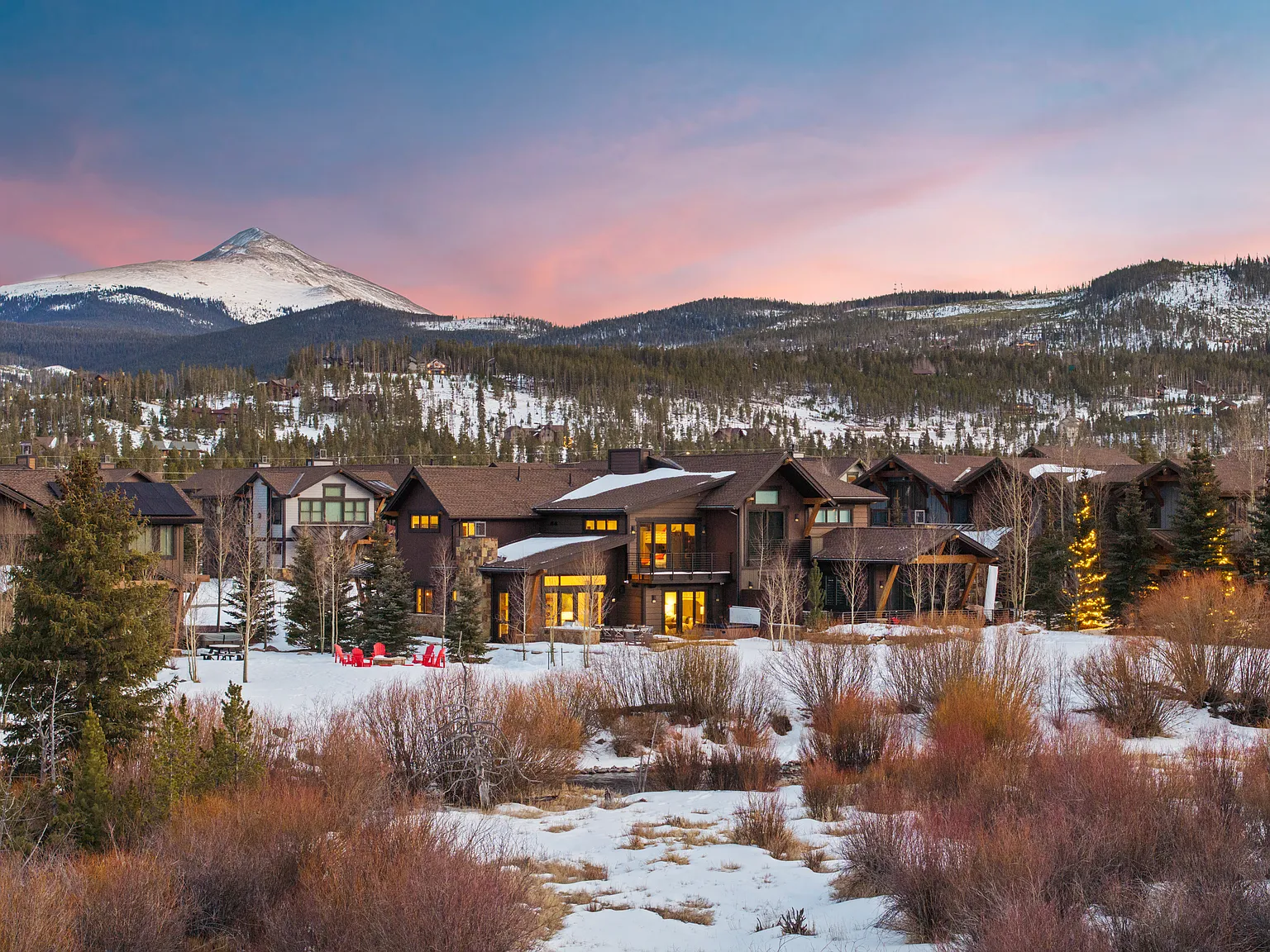 This image showcases the front exterior of a luxurious mountain home during winter. The architecture combines natural stone and wood elements. The house is surrounded by snow-covered landscape with trees and a mountain in the background, evoking a sense of tranquility.