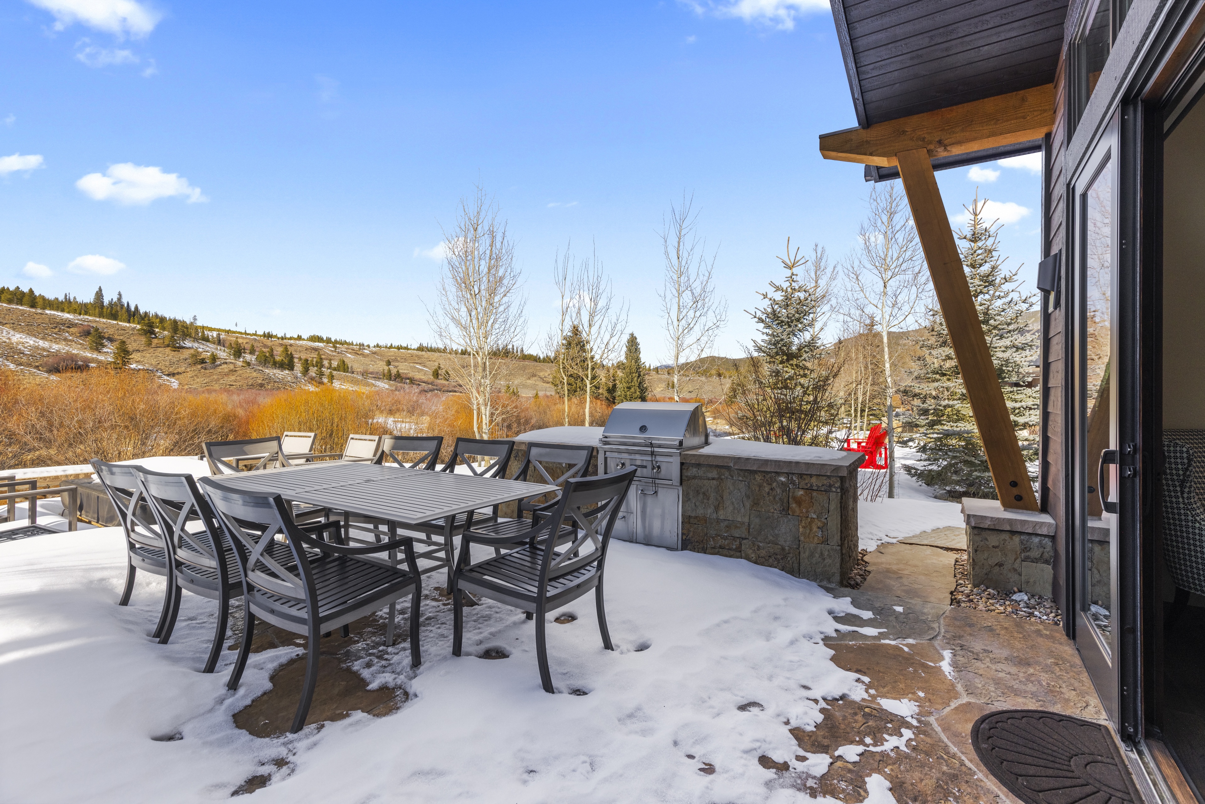 This outdoor patio area offers a beautiful space for dining and relaxing. It features a large table with chairs, a stone-clad outdoor grill station, and a well-maintained stone patio area. A sliding glass door provides access to the interior of the home, complemented by scenic views of the surrounding landscape.
