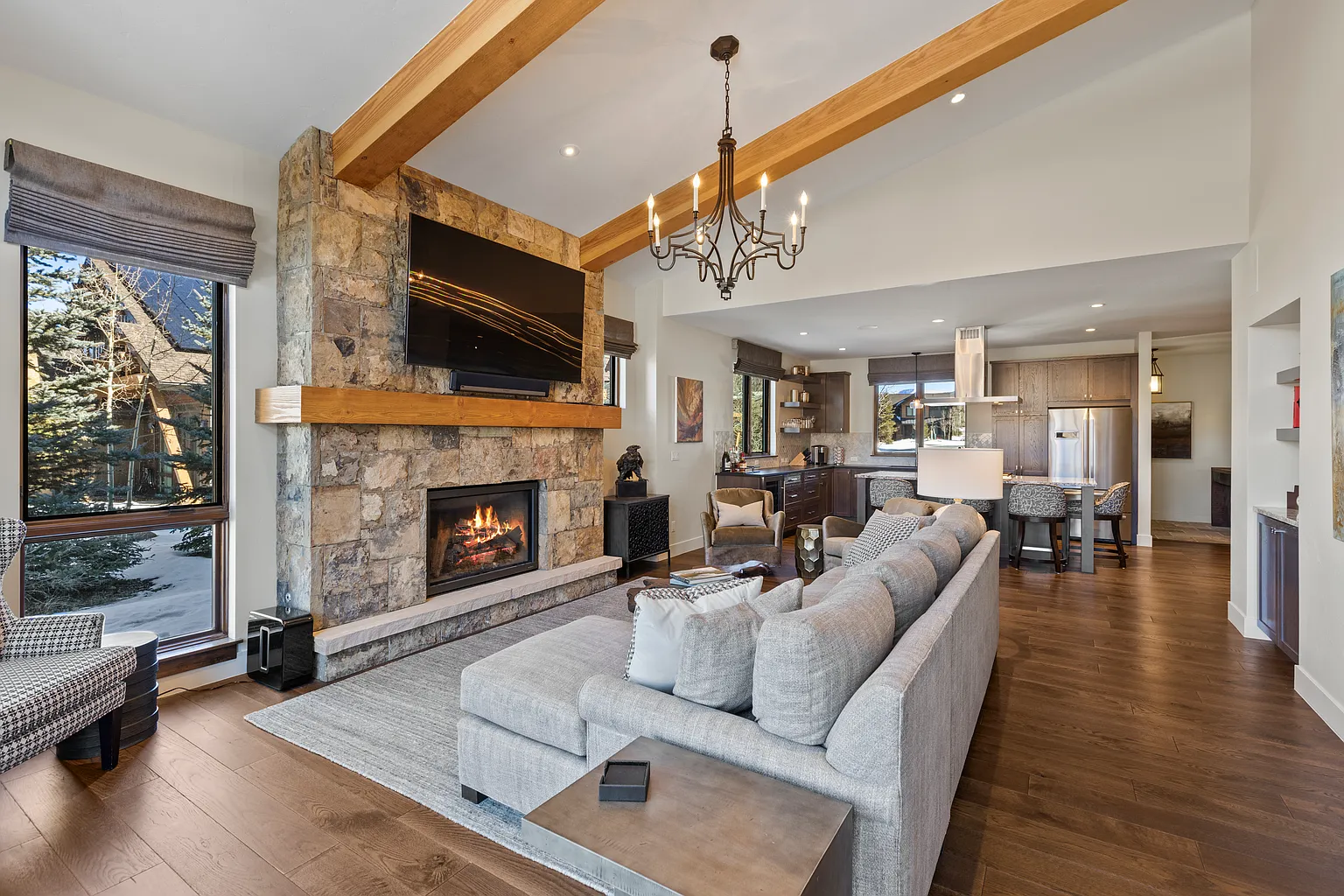 The living room showcases a stone fireplace with a mounted TV, complemented by exposed wooden beams on the ceiling. A large gray sectional sofa anchors the space, while a floor-to-ceiling window allows natural light to flood in. The open-concept design provides a seamless transition to the kitchen, creating a spacious and inviting area.
