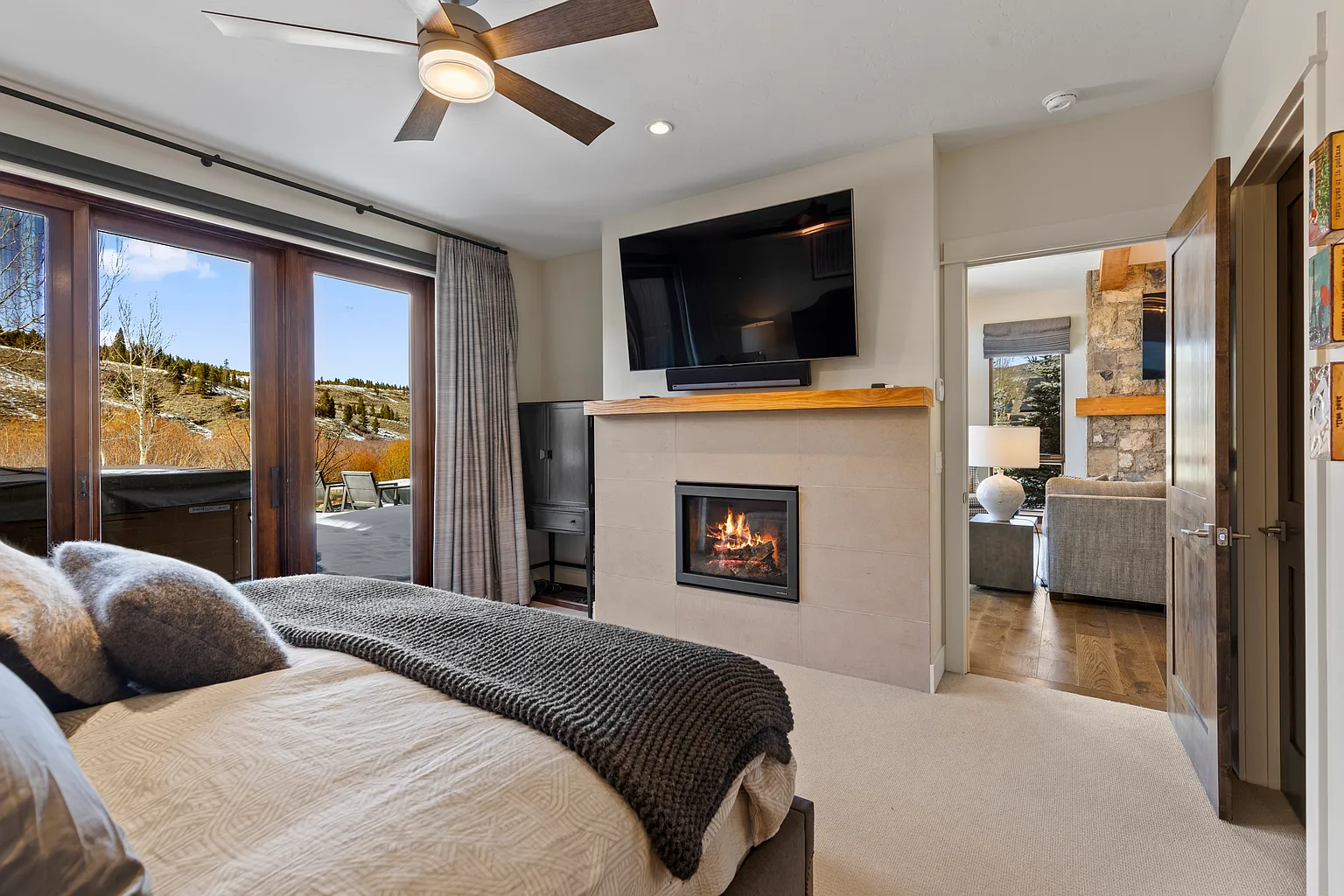 This primary bedroom features a comfortable bed with plush pillows and a cozy throw blanket. A modern fireplace with a mounted television above it adds a luxurious touch to the room. Sliding glass doors lead to a patio/deck area with beautiful outdoor views, enhancing the light and airy atmosphere of the bedroom.