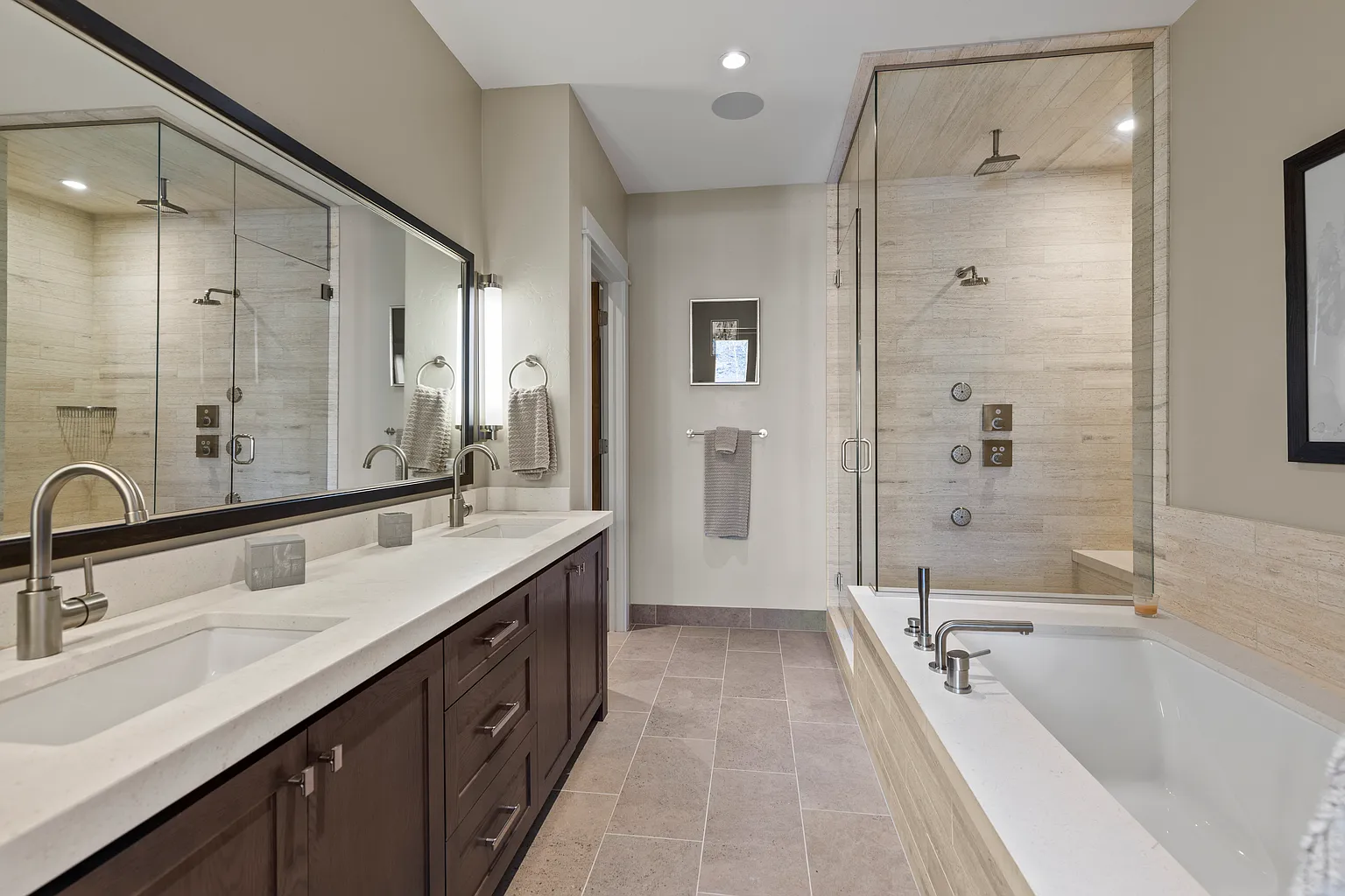 This is a luxurious primary bathroom featuring a double vanity with a white countertop, dark wood cabinetry, and sleek, modern faucets. A large mirror stretches across the vanity area, reflecting the spacious shower and bathtub combination situated behind a glass enclosure. The bathroom is finished with neutral-toned tile flooring, creating a serene and sophisticated ambiance.