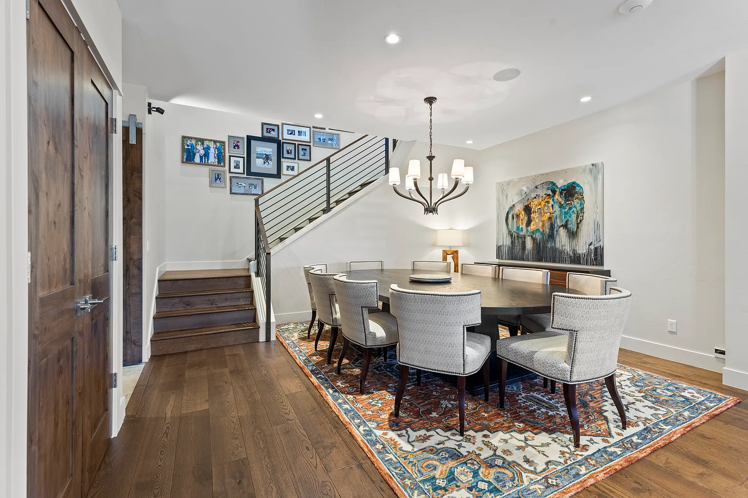 This is a dining room featuring a large dark wood table surrounded by upholstered chairs with dark wood legs. A patterned rug is underneath the table. There's a staircase adjacent to the dining area and artwork hanging on the wall. A chandelier provides light. The overall impression is elegant and inviting.