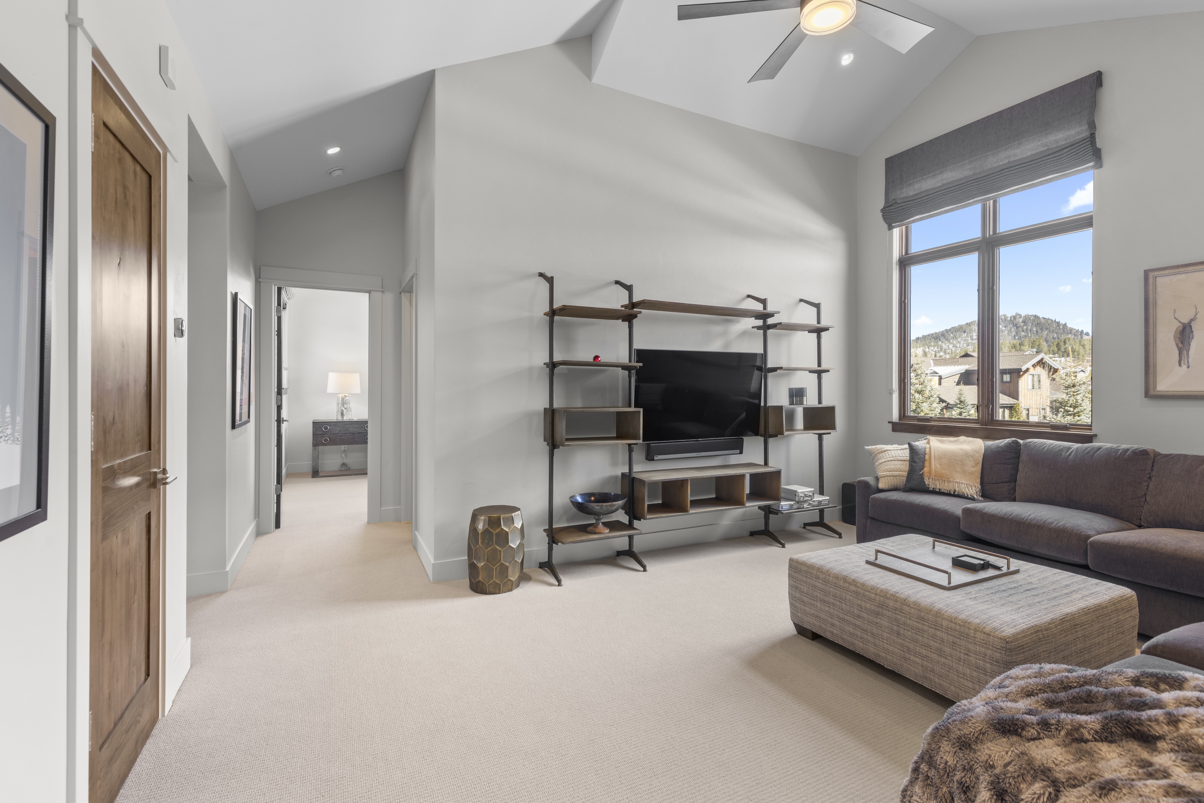 This inviting living room features a modern aesthetic with vaulted ceilings and a large window offering natural light and views. The space is furnished with a comfortable gray sectional sofa, an upholstered ottoman, and a unique shelving unit holding a flat-screen TV. The neutral color palette and plush carpeting create a warm and sophisticated ambiance.