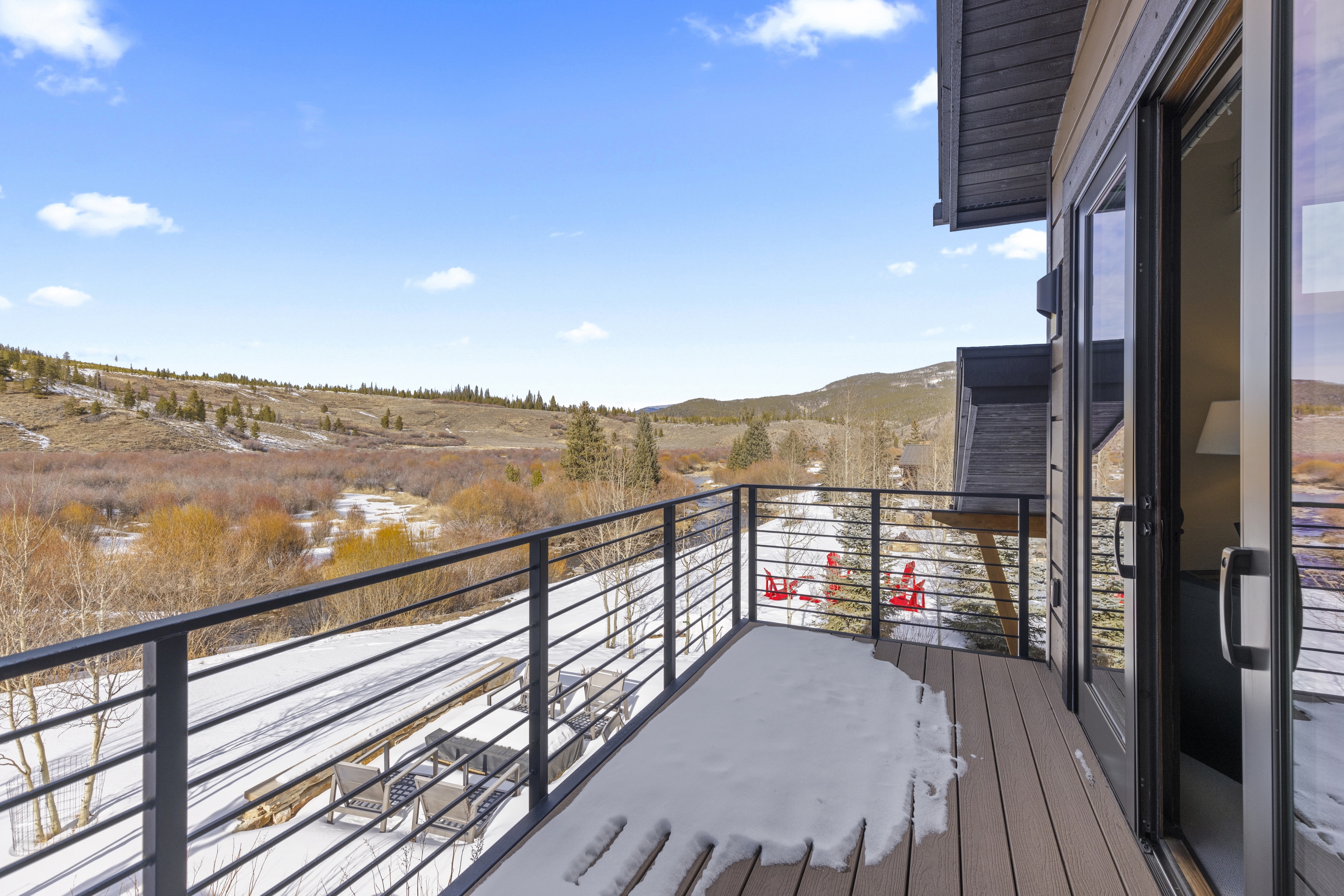 This image showcases a modern balcony with a sleek metal railing, wooden decking partially covered in snow, and a view of a serene landscape with trees and open fields. The balcony is accessible through large sliding glass doors, enhancing indoor-outdoor living. Some outdoor seating or lounging furniture is visible as well as some red chairs in the background.