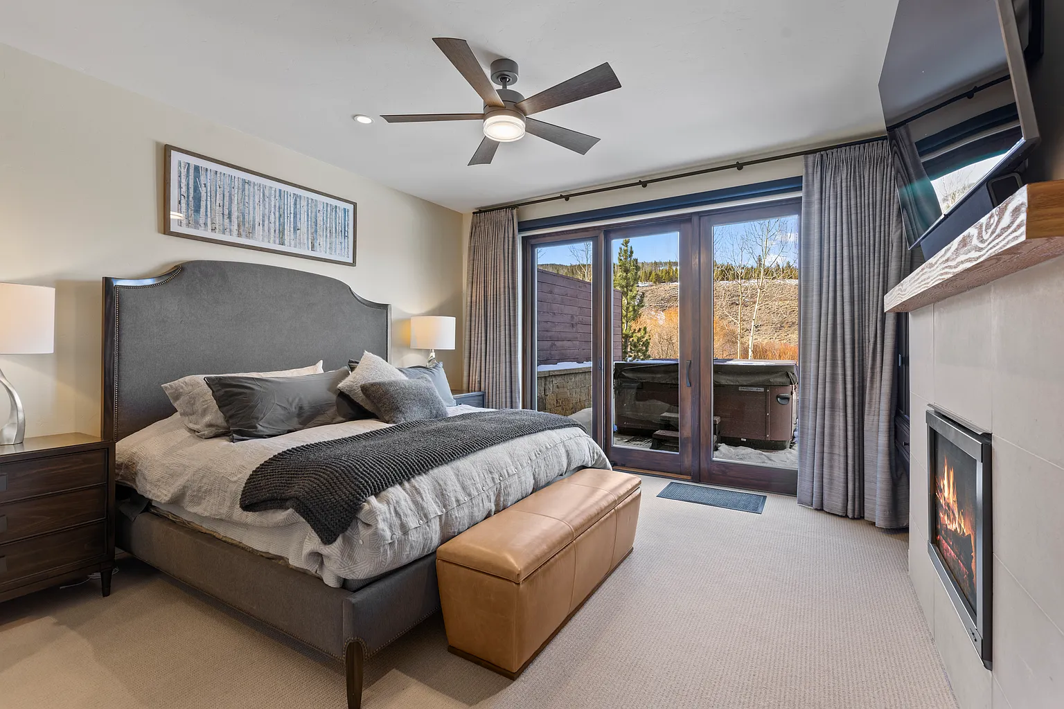 This is a well-appointed primary bedroom featuring a large bed with a gray upholstered headboard, complemented by a neutral color palette and a textured throw. The room has a fireplace and access to a balcony with a hot tub. Overall, the bedroom has a relaxing and luxurious feel, perfect for a real estate listing.