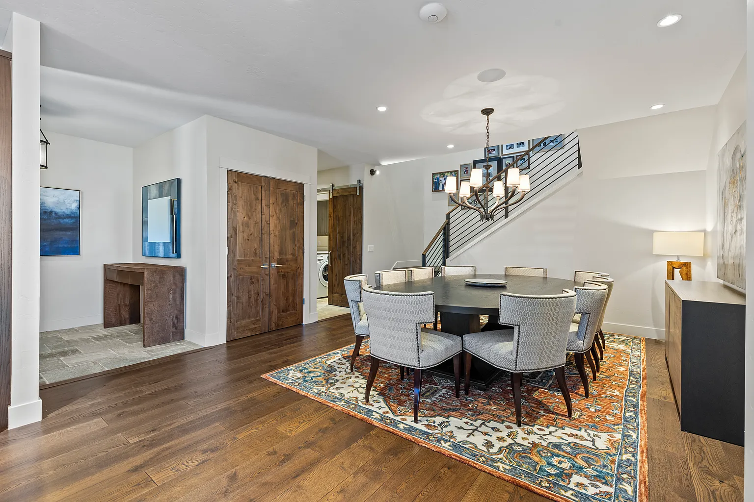 The image showcases a stylish dining room featuring a round dining table with upholstered chairs, set atop a patterned rug that adds visual interest. Wood flooring and natural-toned walls create a warm ambiance, while modern lighting fixtures and a staircase in the background add architectural and design appeal, making it an inviting space for meals and gatherings. A sideboard and artwork are also visible.