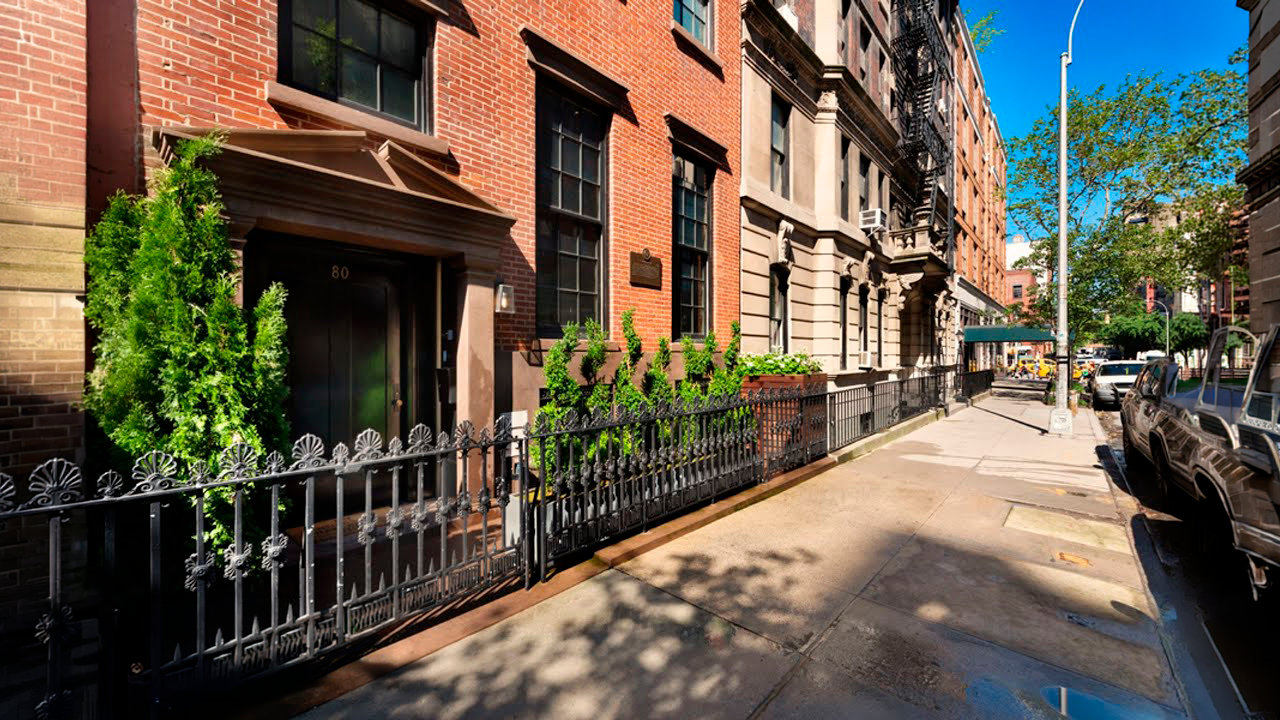 The image captures the front exterior of a classic brownstone-style building. A wrought iron fence surrounds a small garden area in front of the building. The facade features a red brick exterior with dark-framed windows and an elegant entryway, contributing to a stately and sophisticated street presence.