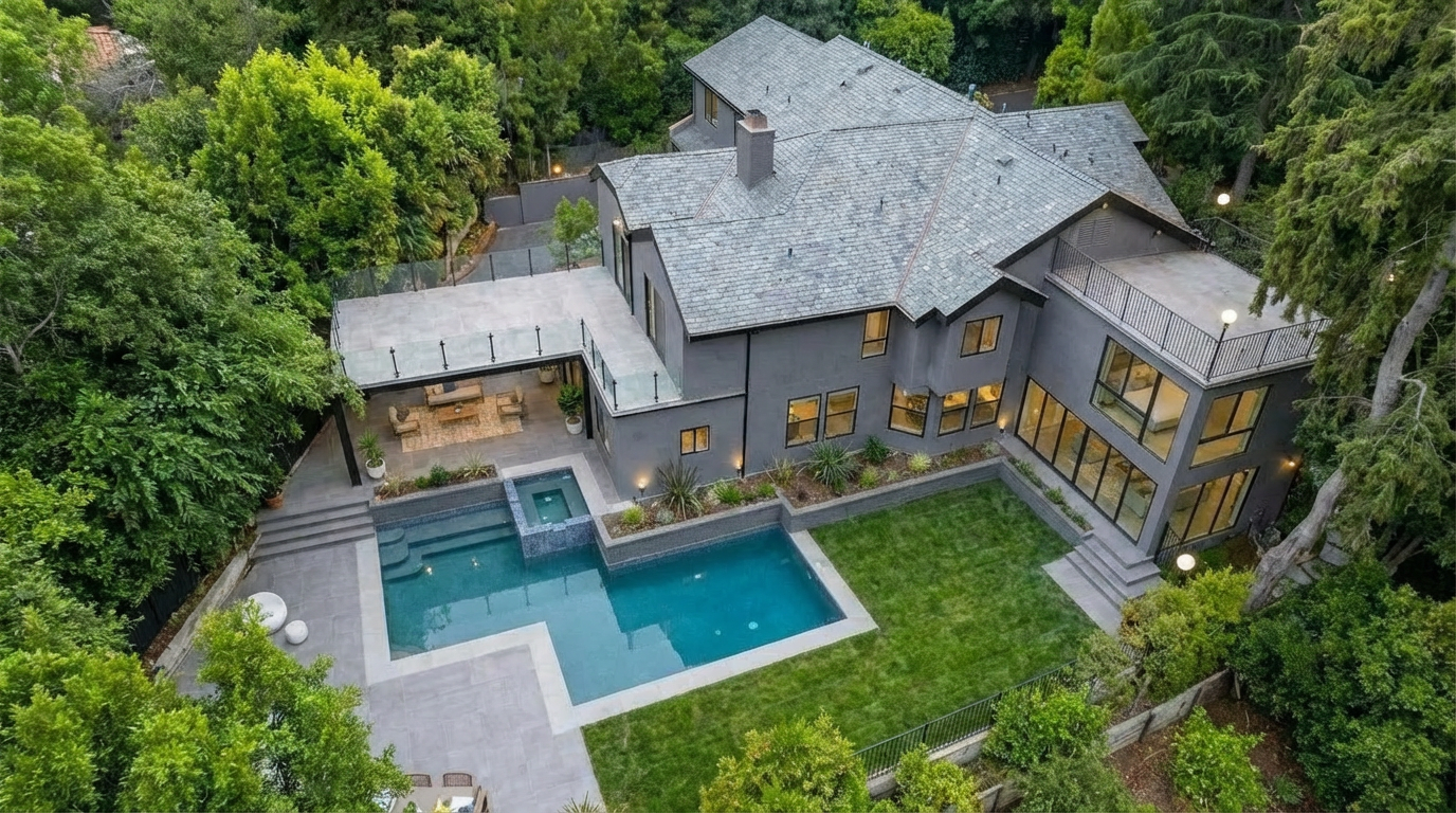 This aerial view showcases a luxurious modern home surrounded by lush greenery. A prominent feature is the backyard with a large swimming pool, a spa, and a well-manicured lawn. The main house boasts a gray exterior with a slate roof, multiple windows, and an attached patio area, presenting an impressive and private outdoor living space.