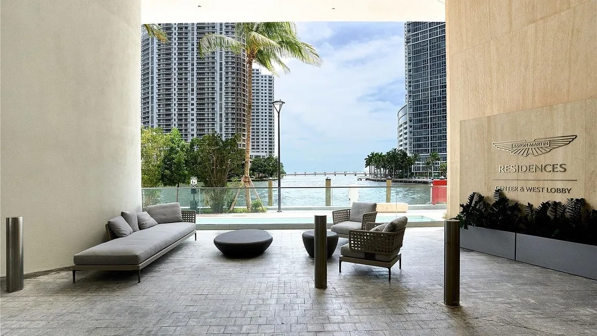This image showcases an open-air patio or balcony area with contemporary outdoor furniture, including a chaise lounge and armchairs. The area overlooks a waterfront view with high-rise buildings in the background, offering a luxurious and relaxing ambiance. The Aston Martin Residences logo on the wall indicates a high-end property.