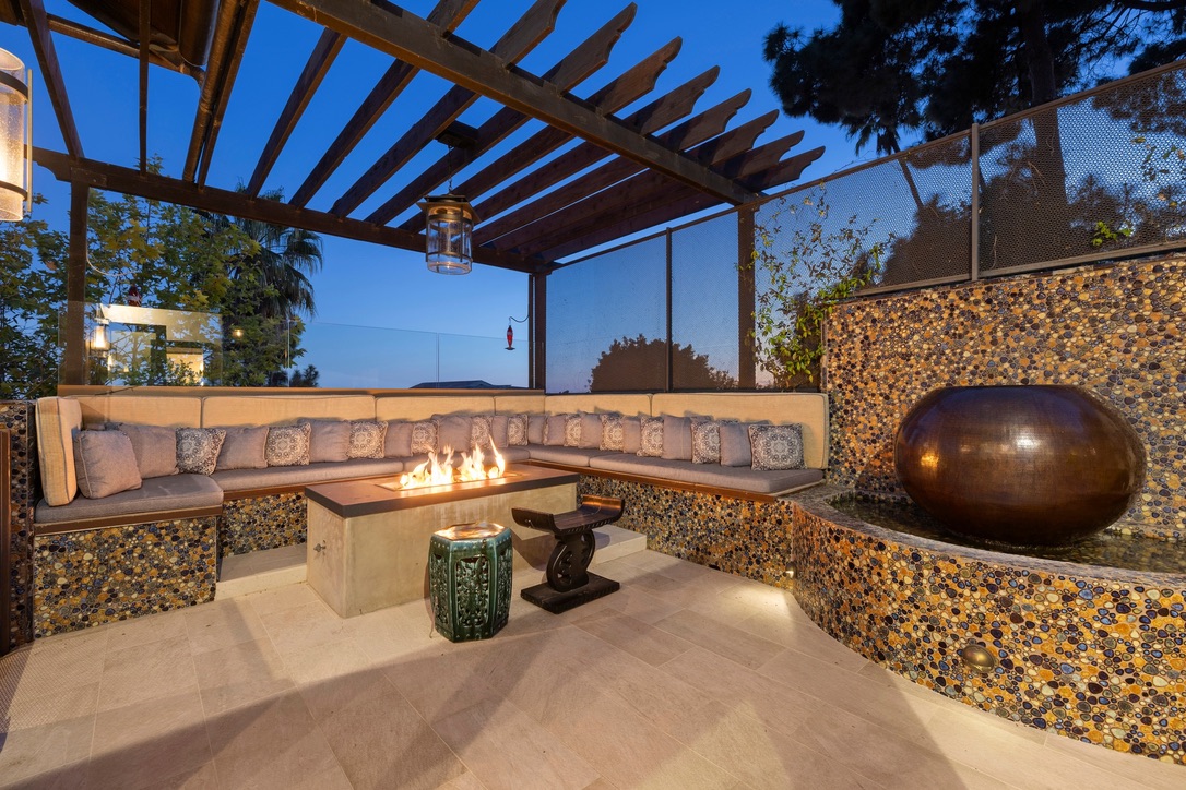This inviting outdoor lounge area features a built-in L-shaped bench with plush seating and numerous patterned throw pillows, centered around a rectangular fire pit. The space is defined by a wooden pergola overhead and a unique, textured stone-tiled wall that incorporates a large, spherical water feature. The scene is captured at dusk, highlighting the warm glow of the fire and integrated landscape lighting, creating a sophisticated and cozy atmosphere for evening relaxation.
