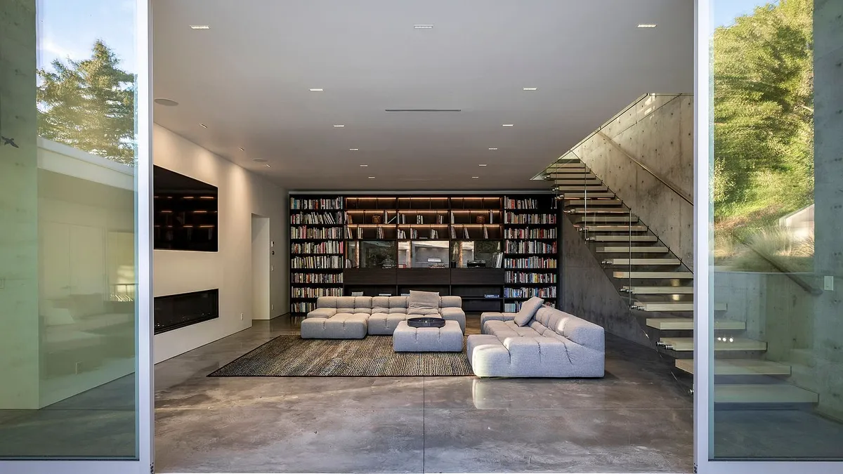 This is a modern living room featuring a large modular sofa, a floor-to-ceiling bookshelf, and a sleek fireplace. A contemporary staircase leads to the upper level, adding an architectural element. The space is well-lit and has a polished concrete floor that enhances the minimalist aesthetic.