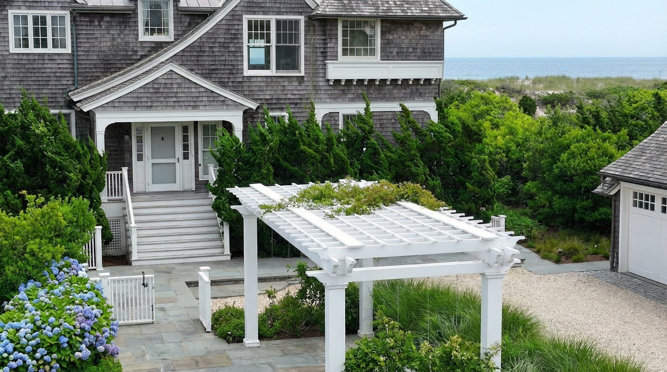 This image showcases the front exterior of a charming home, likely a beach house, with a shingled facade and classic architectural details. A white pergola, lush greenery, and hydrangea bushes add to the property's curb appeal. The view hints at a coastal location, making this property an attractive residence.