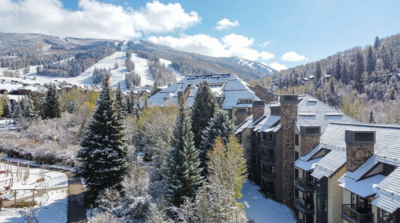 This aerial shot showcases a snow-dusted mountain resort, highlighting both residential buildings and ski slopes. The architecture features multi-story buildings with stone and timber accents, suggesting a luxurious mountain retreat. The scene conveys a sense of winter serenity and premium living in a scenic location.