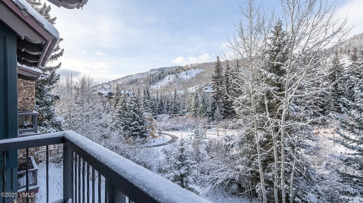 This image showcases a snowy balcony view, emphasizing the scenic winter landscape. The surrounding area is blanketed in snow-covered trees, with a path winding through a park-like setting visible below. In the distance, snow-covered hills add depth and appeal, suggesting a serene and picturesque mountain retreat.