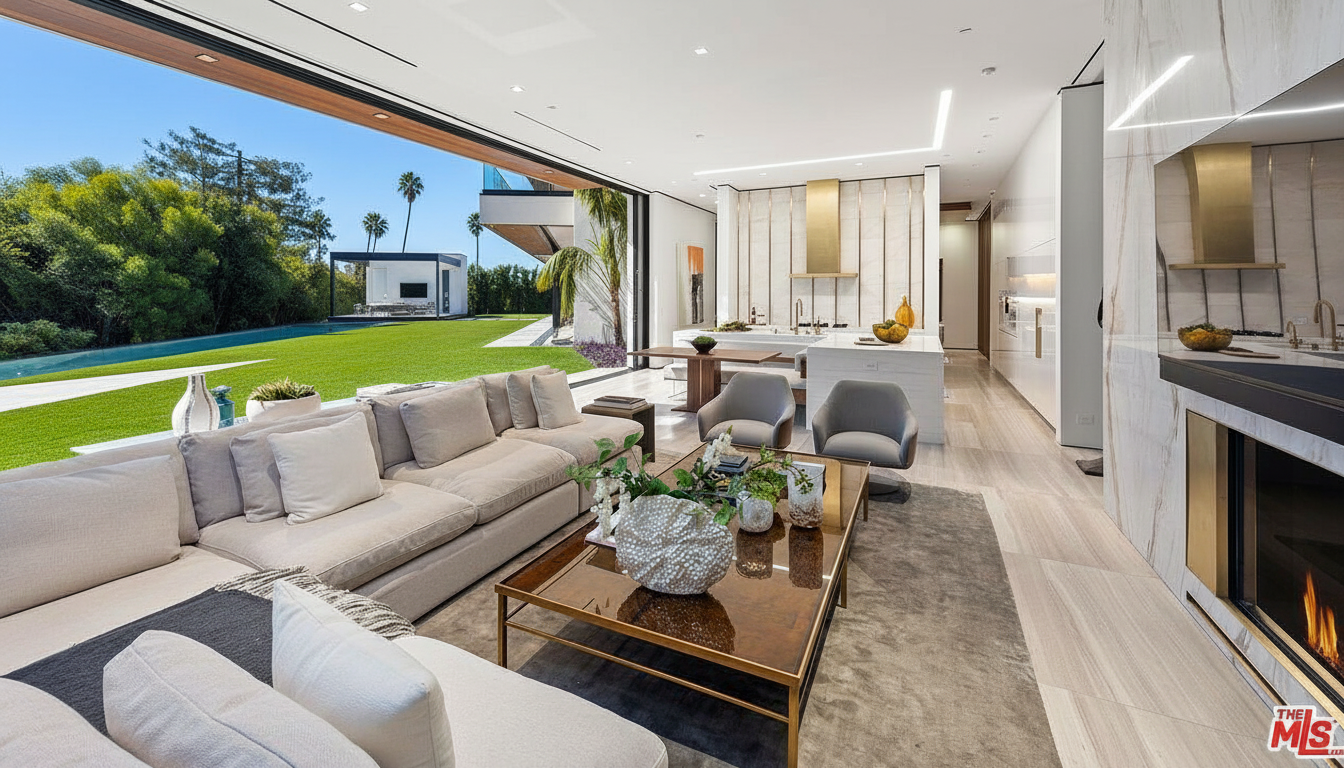 This interior showcases a modern living room seamlessly blending with outdoor living. A large sectional sofa centers the space, facing a cozy fireplace. The open floor plan extends to a sleek kitchen and dining area, all while overlooking a well-manicured lawn, pool, and a cabana, creating a luxurious indoor-outdoor living experience.