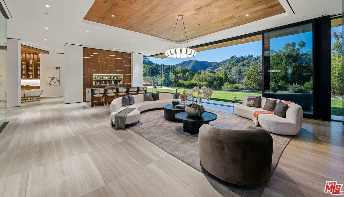 This luxurious living room boasts a modern design with curved sofas, dark wood accents, and floor-to-ceiling windows offering scenic views. A stylish chandelier hangs above a central seating area with round coffee tables and a plush rug. The open layout and high-end finishes create a sophisticated and inviting space, perfect for entertaining.