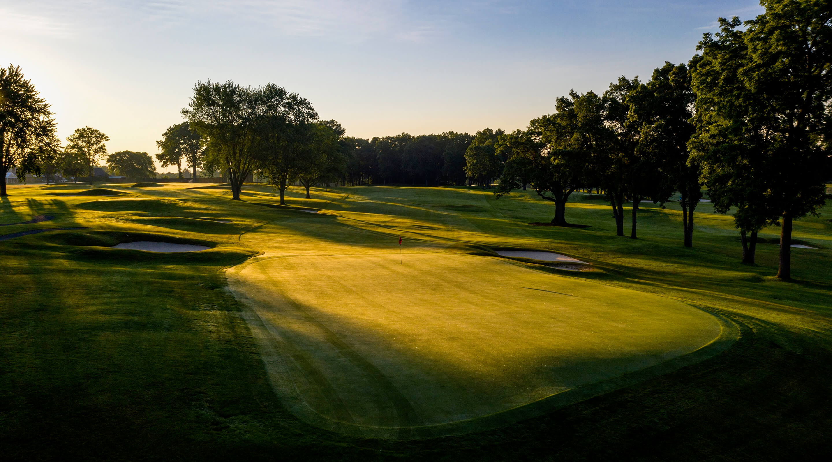 This aerial shot showcases a beautifully manicured golf course at sunrise or sunset. The green is vibrant and well-maintained, with mature trees lining the fairways and strategic bunkers in place. The overall impression is one of luxury and tranquility, highlighting a desirable recreational amenity.