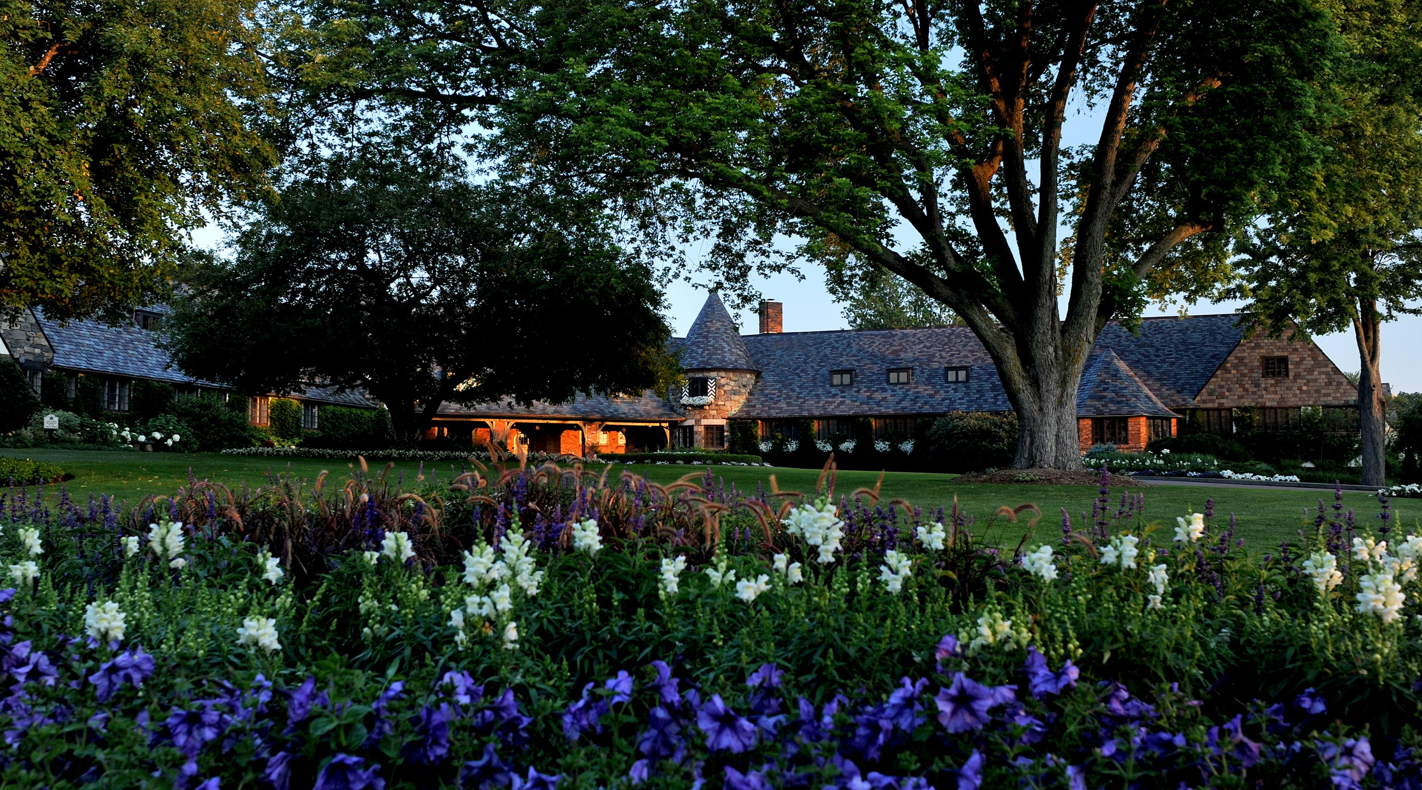 This image showcases the exterior of a grand estate, focusing on its meticulously landscaped yard and garden. A diverse array of flowers in shades of white and purple provides a foreground of vibrant color, leading the eye to the impressive architecture hidden behind mature trees. The well-maintained lawn and the thoughtful placement of plants suggest a property of considerable size and value.