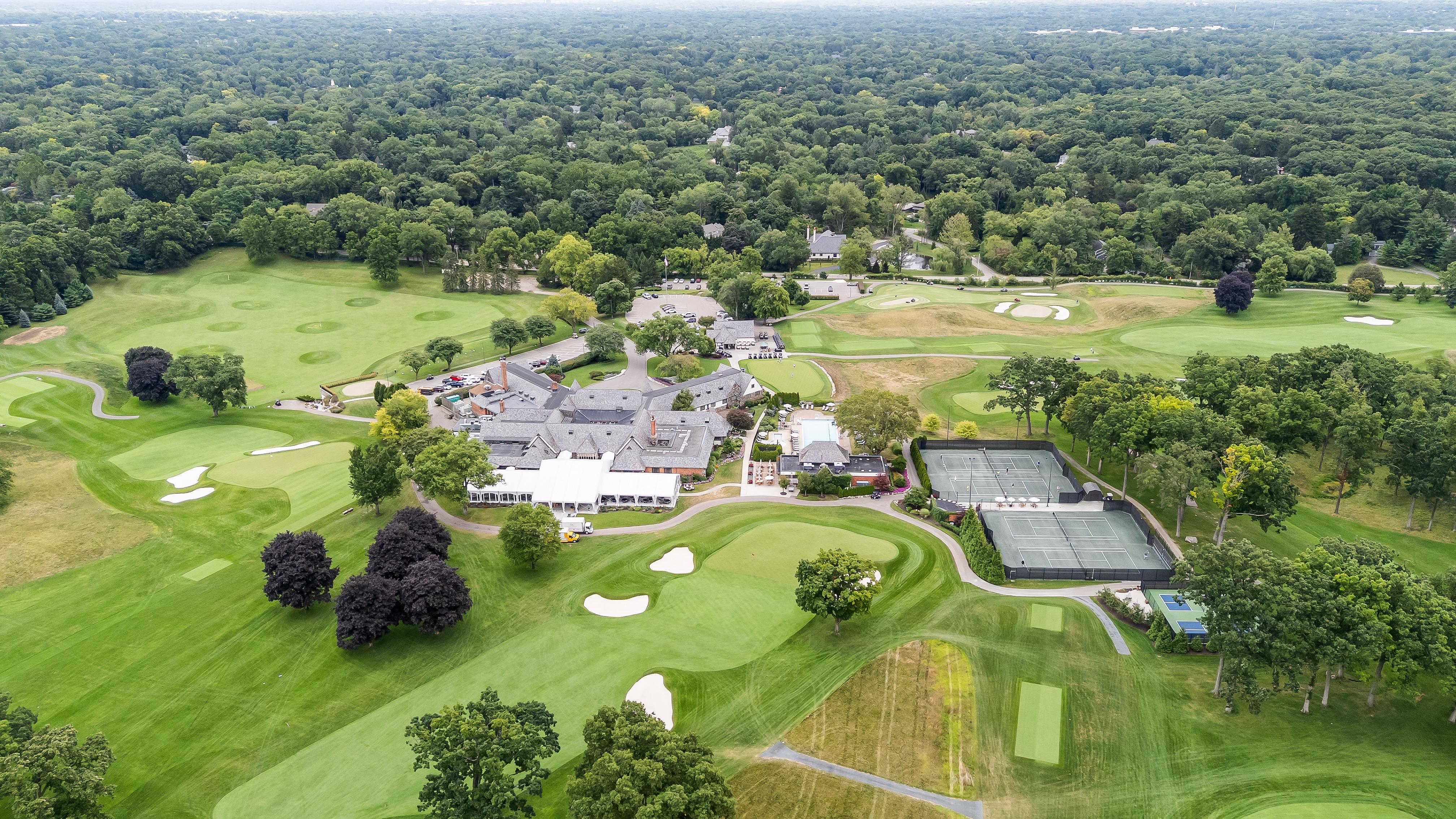 This aerial view showcases a sprawling golf course and country club. The image highlights the meticulously manicured greens, mature trees, tennis courts, and the grand clubhouse with its extensive rooftops. This view emphasizes the expansive property and recreational amenities suitable for luxury real estate marketing.