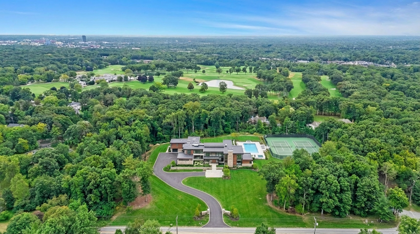 This aerial shot showcases an expansive property featuring a modern house nestled amidst lush greenery. A long driveway leads to the house, which boasts a swimming pool and tennis court in the backyard. In the background, a golf course stretches out with hints of a distant cityscape, creating a luxurious and private atmosphere.
