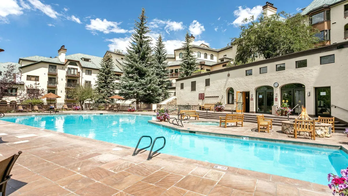 This outdoor pool area serves as a significant community amenity, featuring a crystal-clear swimming pool and an adjacent hot tub. Surrounded by stone decking, a fire pit, and comfortable patio furniture, the space is set against a backdrop of mature pine trees and a multi-story building with a cozy, mountain-resort architectural style. The area is well-maintained and inviting, perfect for relaxation and socializing.