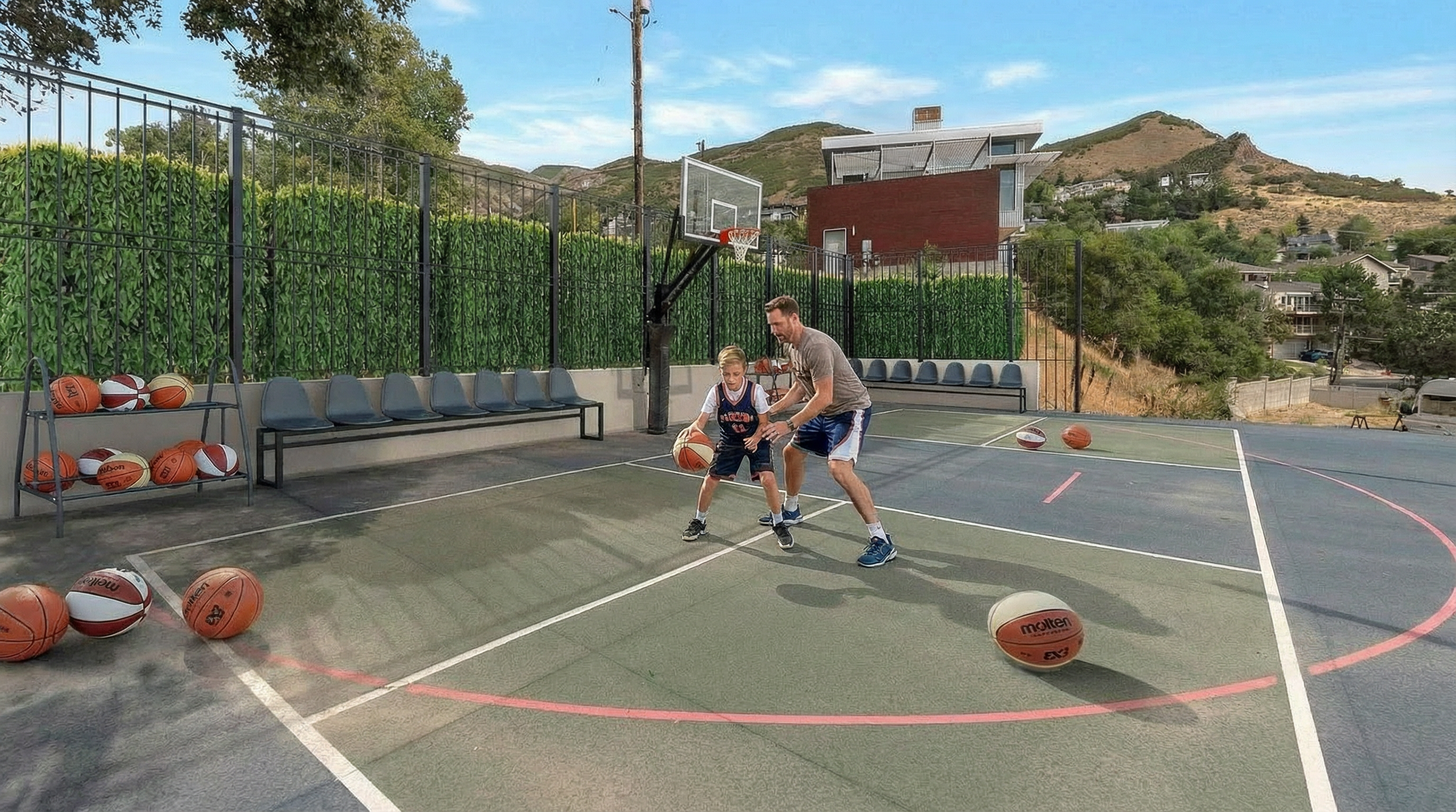 This image showcases a basketball court, likely part of a community amenity or a private residence. A boy and a man are playing basketball on the court. The court includes spectator seating behind a fence, a basketball hoop, and a view of a house and treed hillside in the background.